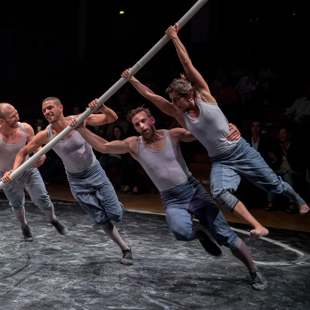 Tanztheater auf der Bühne: Performer balancieren lange Stäbe im Scheinwerferlicht.