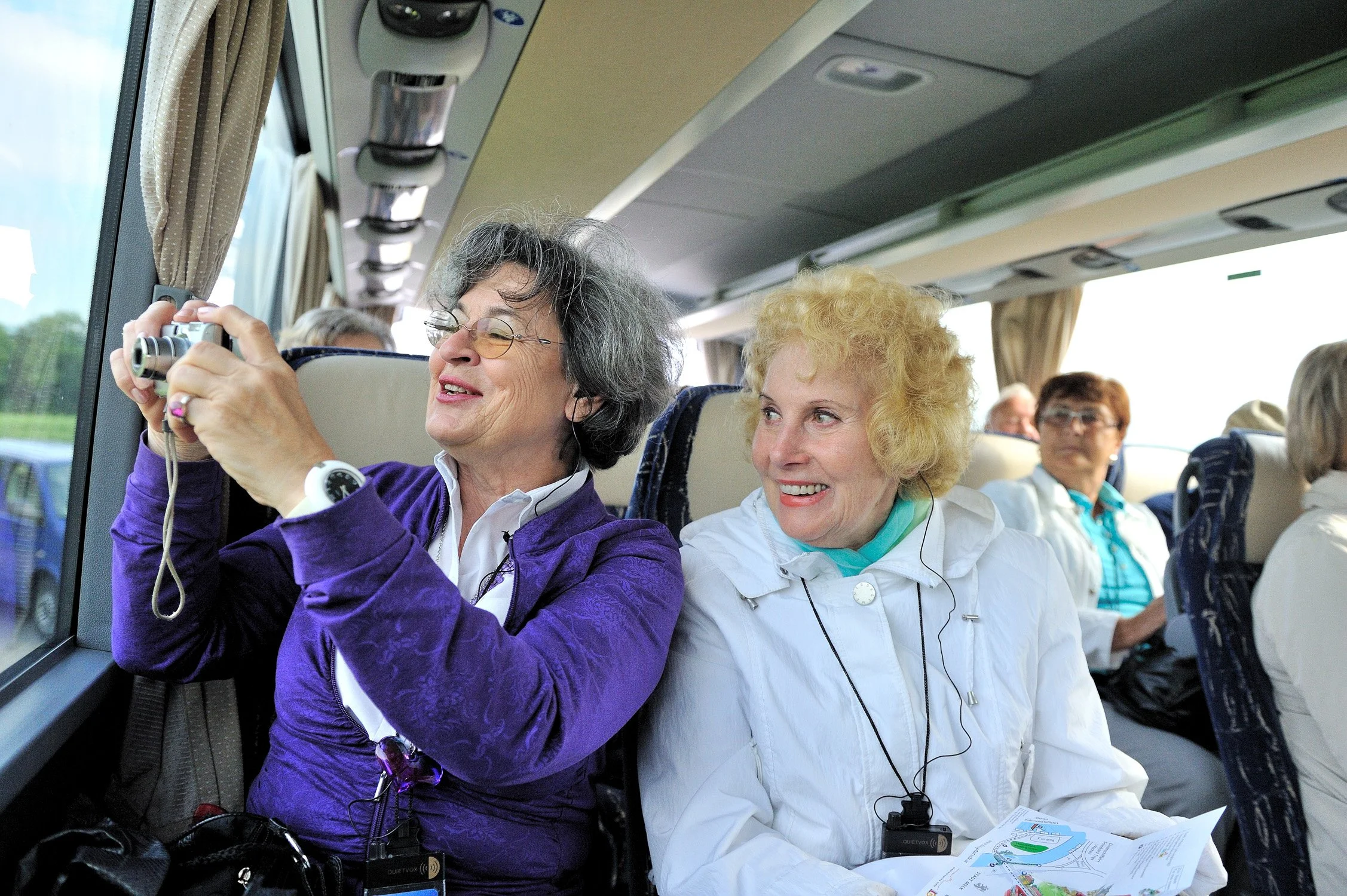 Zwei Seniorinnen im Reisebus, eine fotografiert aus dem Fenster – Reisereportage einer Gruppenreise