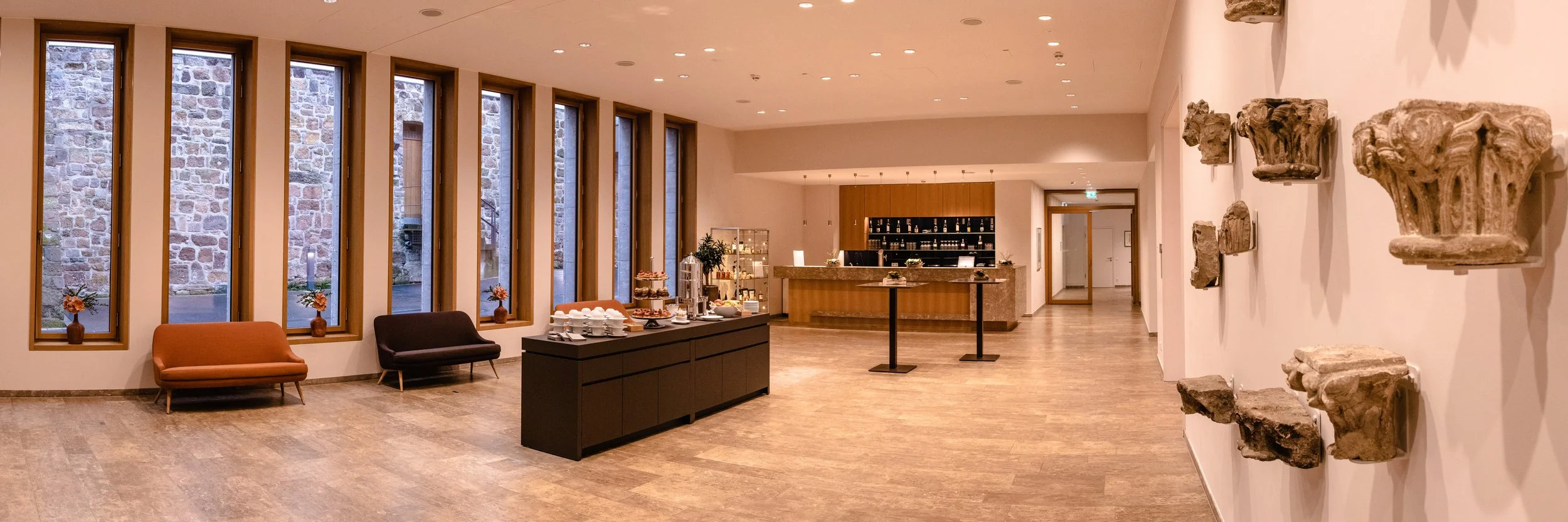 Weiträumiges Foyer mit hohen Fenstern, Empfangstresen und historischen Steinfragmenten an der Wand – Architekturfotografie als Panorama