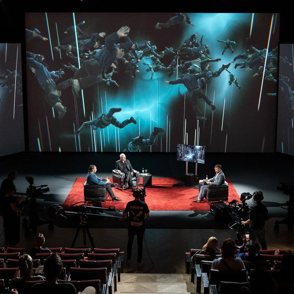 Weite Theaterbühne mit Projektionsleinwand während einer Abendveranstaltung.
