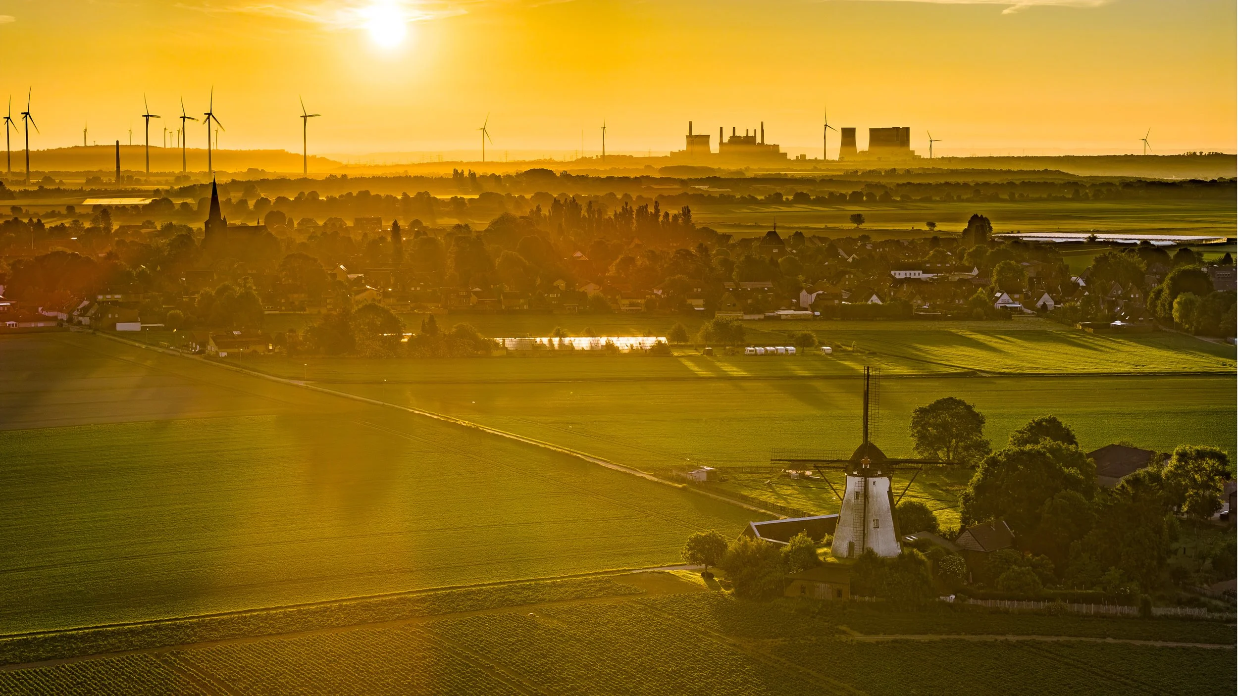 Luftaufnahme einer historischen Windmühle im goldenen Sonnenaufgang, umgeben von Feldern, mit Windrädern am Horizont.