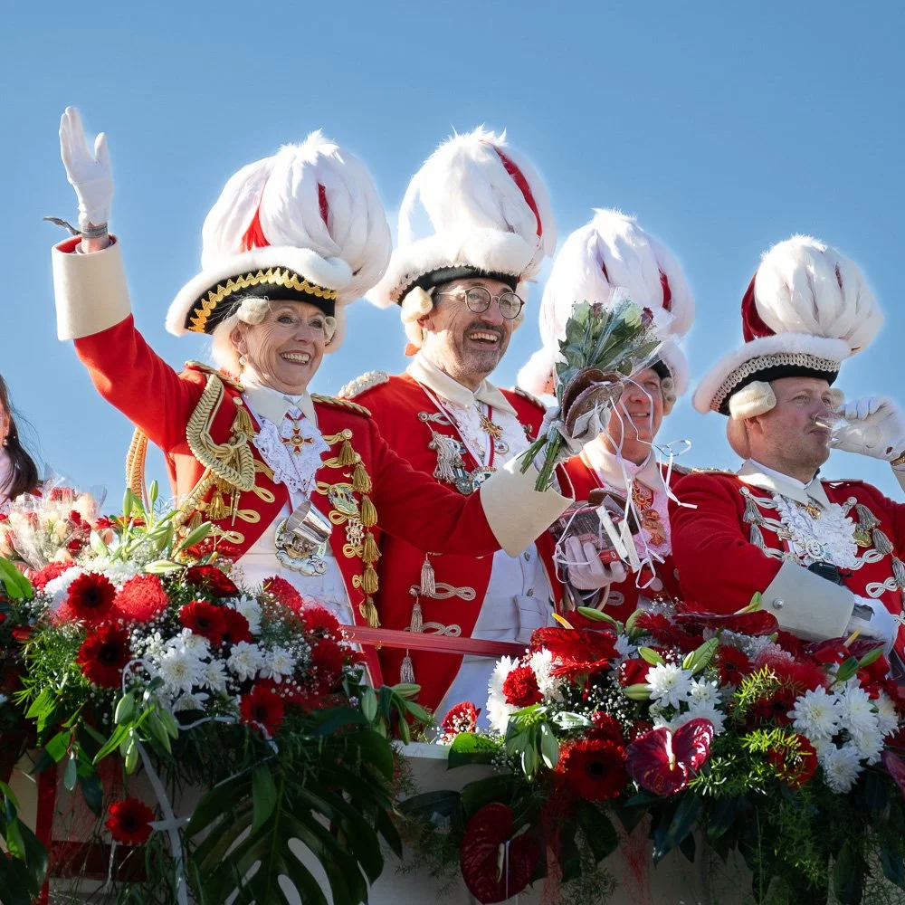 Karnevalsgarde in roter Uniform mit weißen Federhüten unter blauem Himmel.