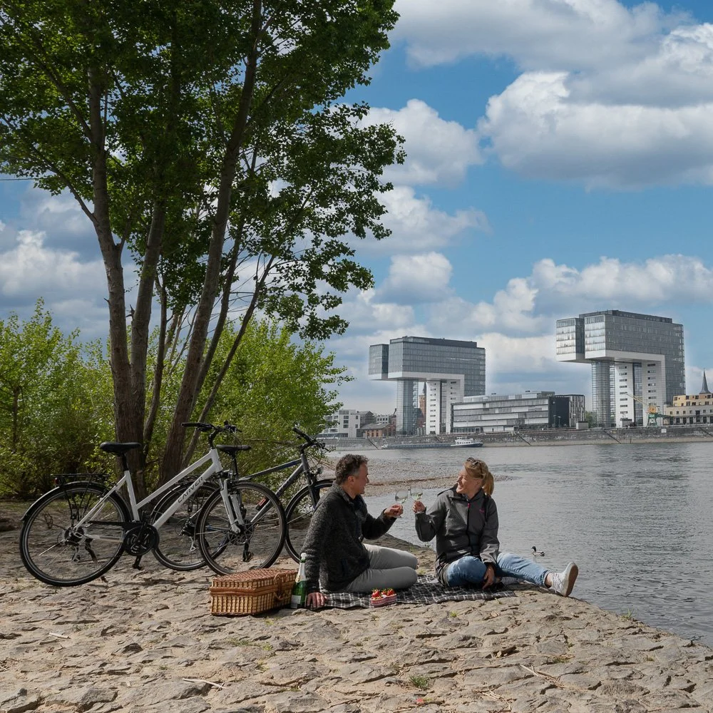 Paar beim Picknick am Rheinufer mit Blick auf die Kölner Kranhäuser, Fahrräder im Vordergrund – Reisefotografie in Köln