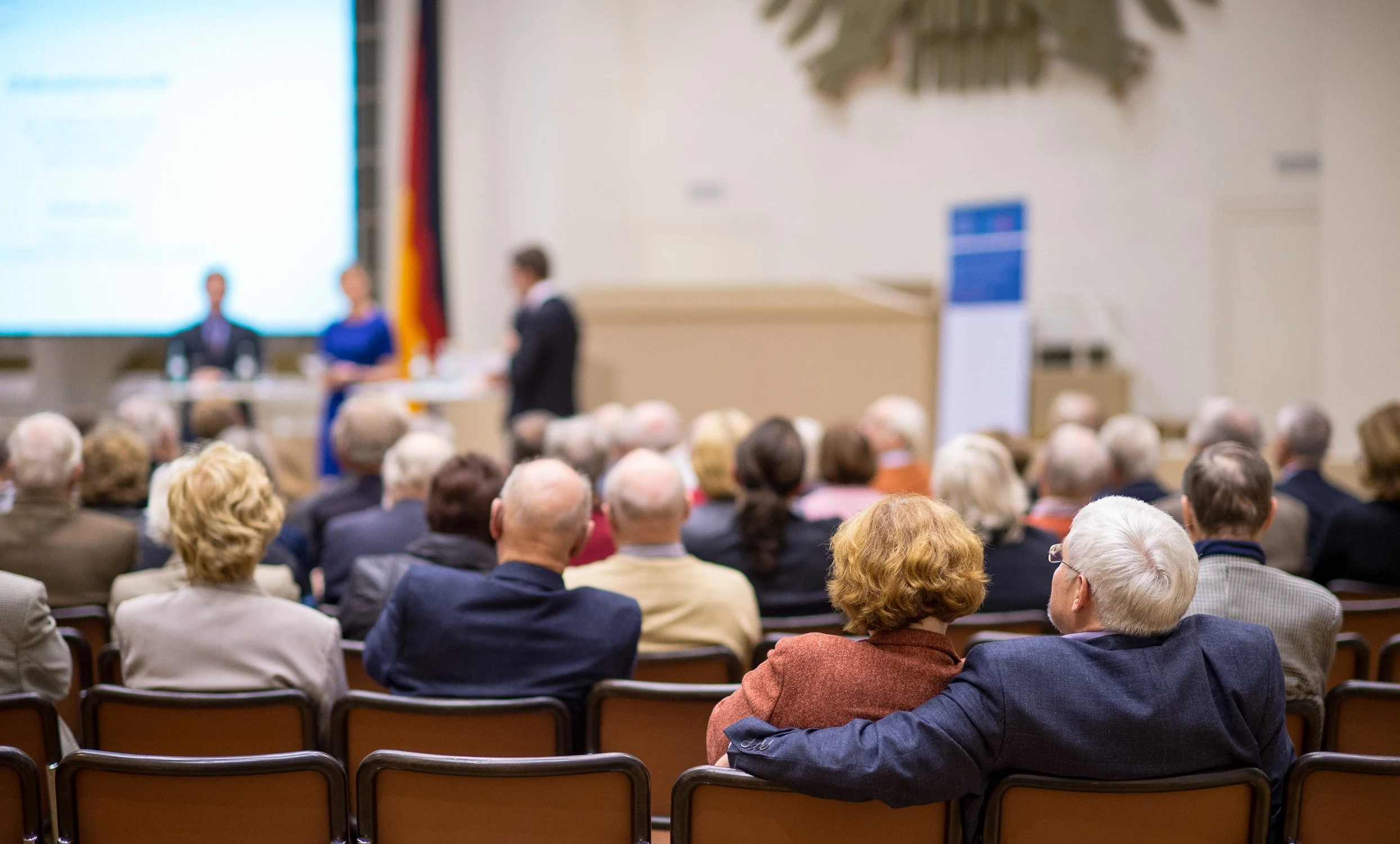 Publikum bei einer politischen Veranstaltung in einem Saal mit Bühne und Leinwand.