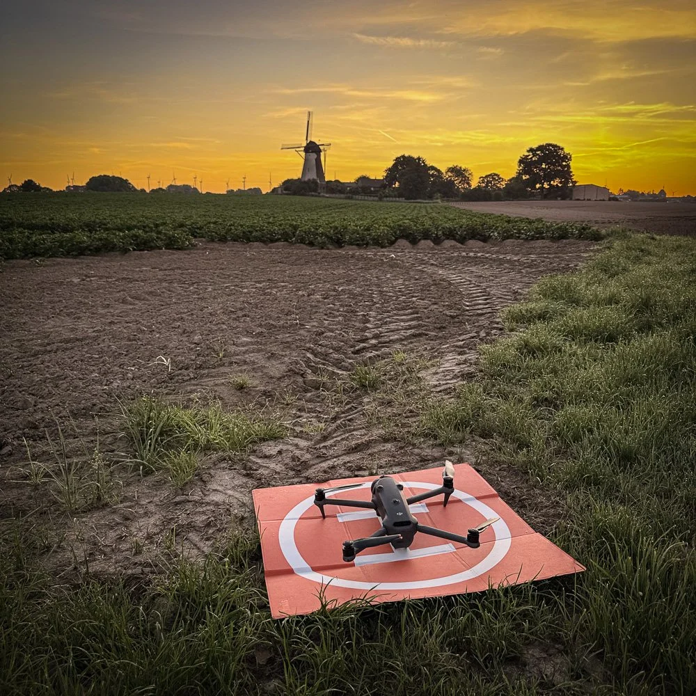 DJI-Drohne auf orangefarbenem Landepad am Feldrand, Windmühle und Felder im Abendlicht des Sonnenuntergangs.