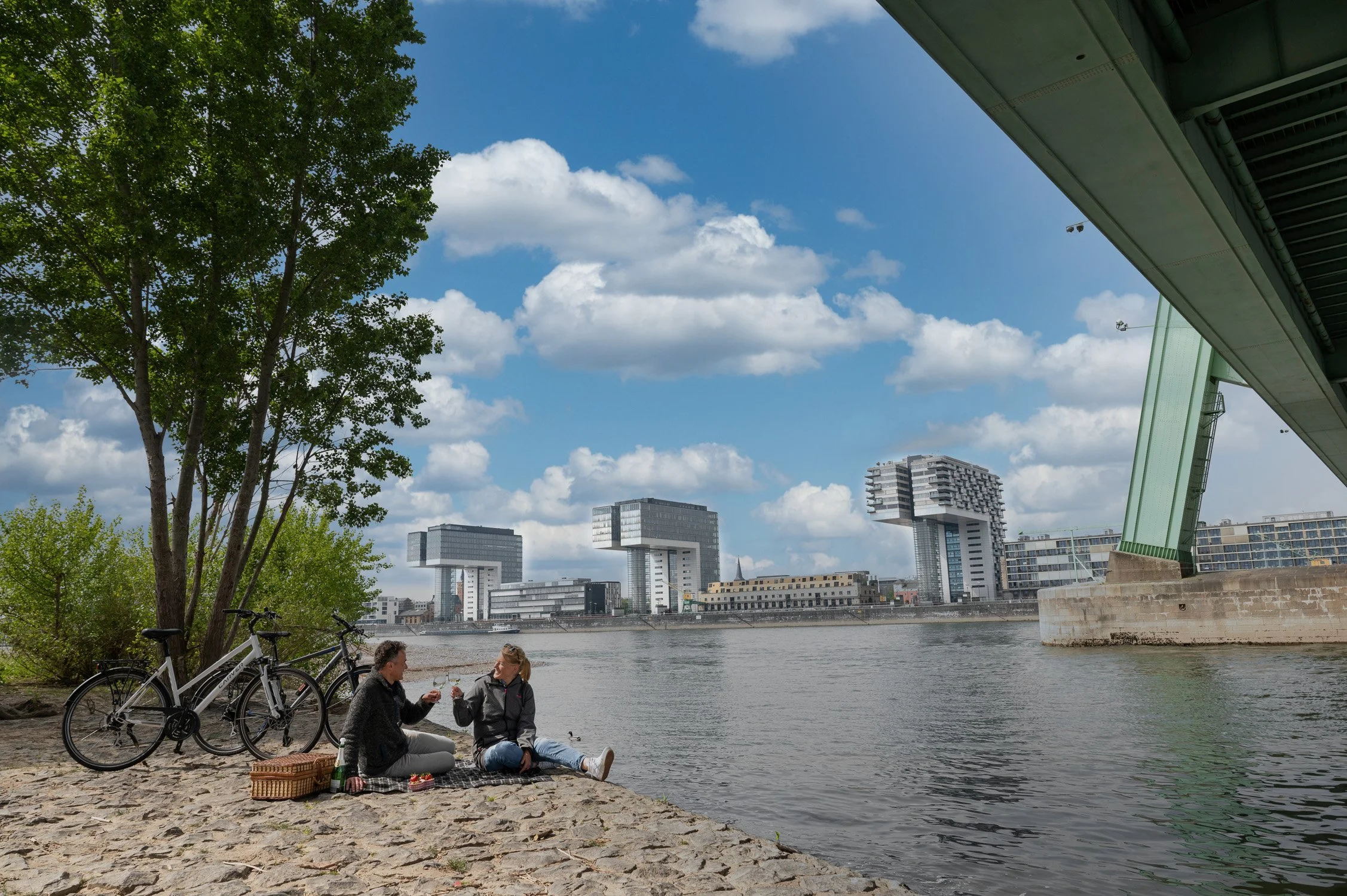 Paar beim Picknick am Rheinufer mit Blick auf die Kölner Kranhäuser, Fahrräder im Vordergrund – Reisefotografie in Köln