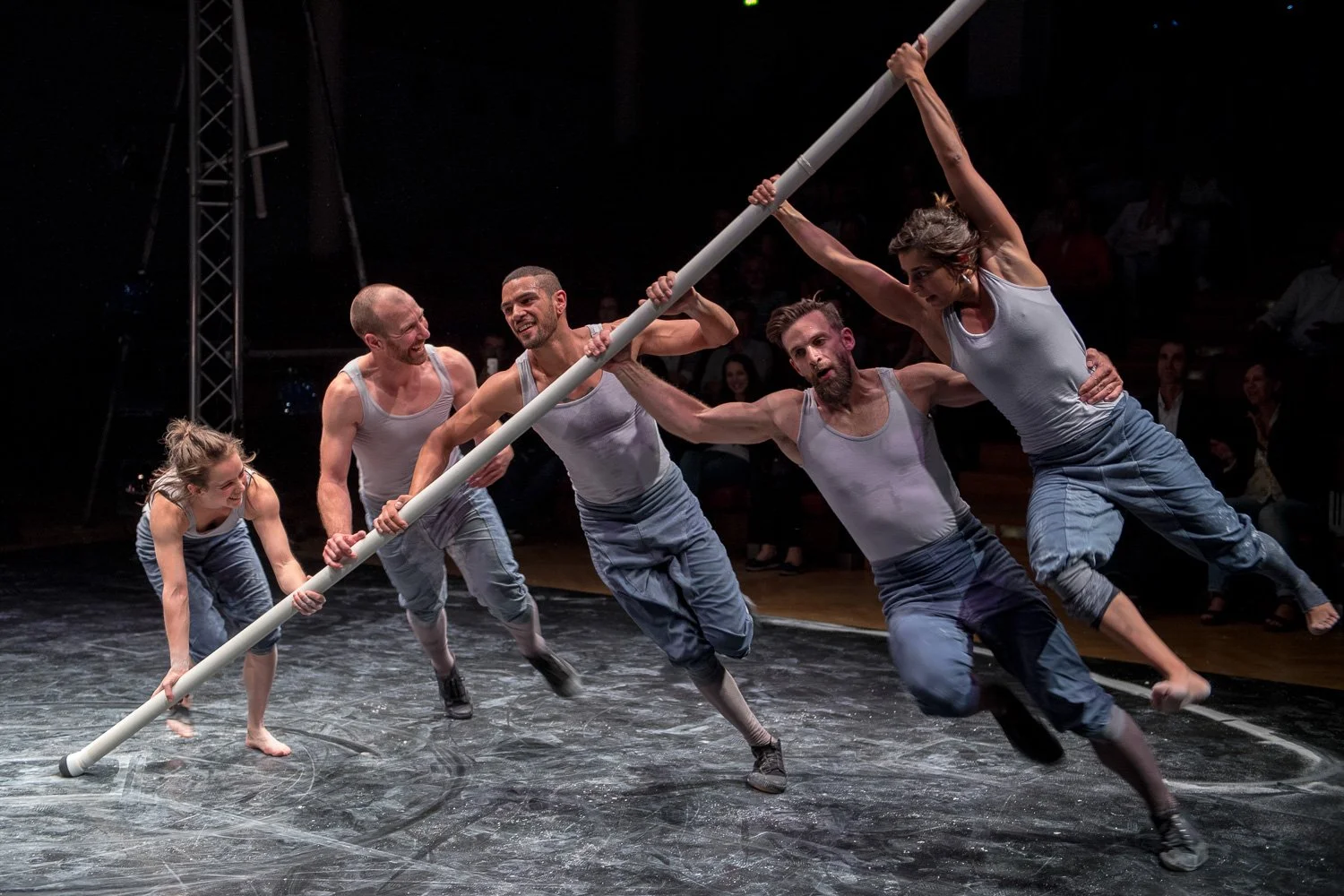 Tanztheater auf der Bühne: Performer balancieren lange Stäbe im Scheinwerferlicht.