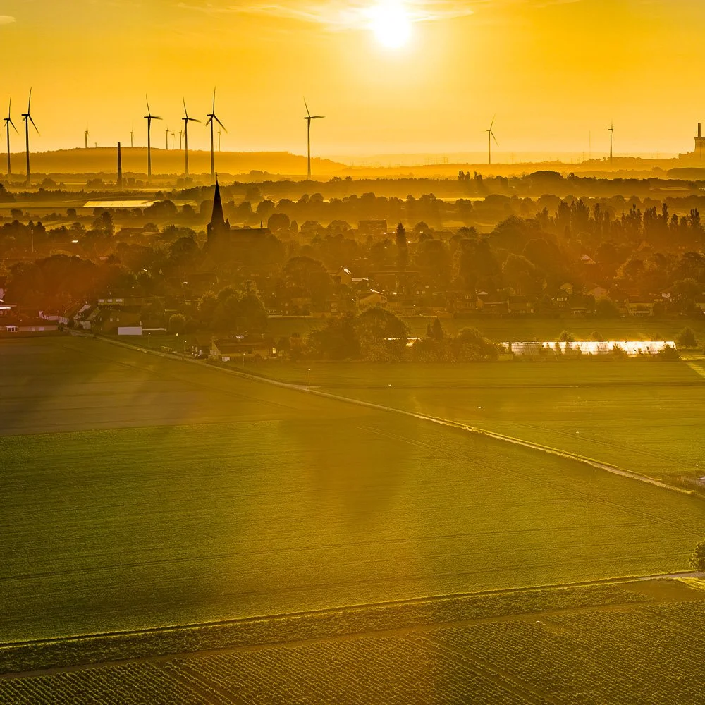 Luftaufnahme im goldenen Sonnenaufgang: Felder, ein Dorf mit Kirchturm und Windräder am dunstigen Horizont.