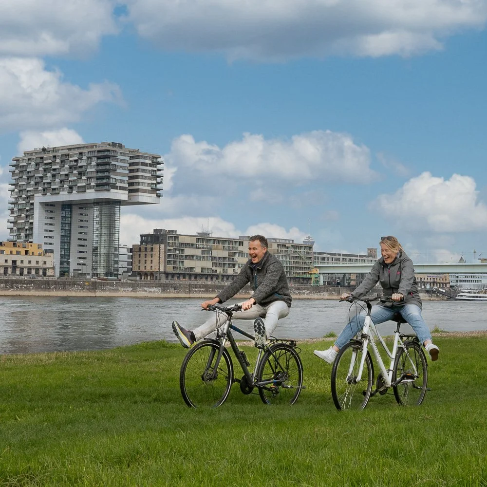 Radfahrer-Paar fährt auf der Rheinwiese vor den Kölner Kranhäusern – Tourismusfotografie in Köln