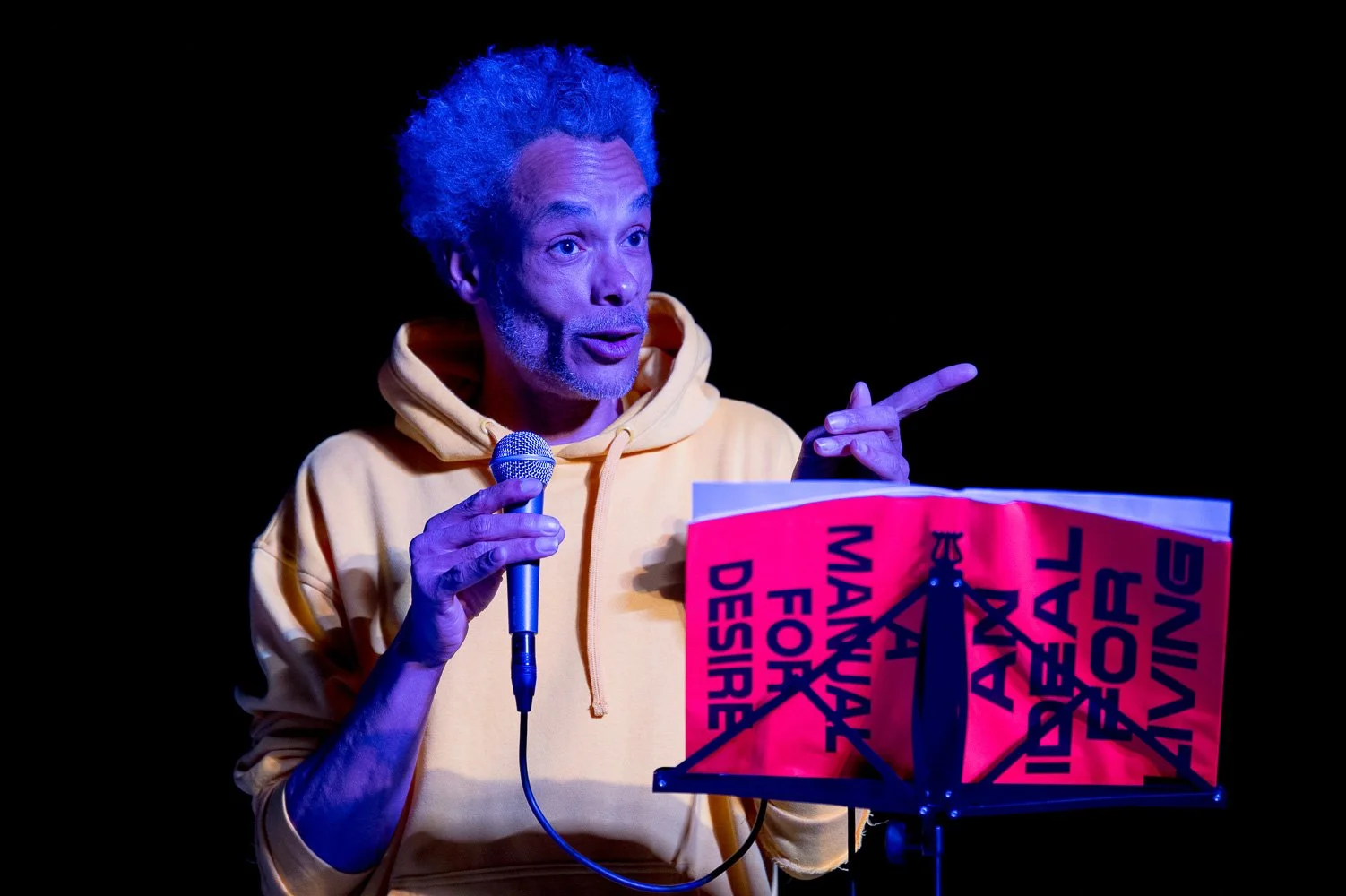 Performance-Künstler am Pult mit Schild in blauem Bühnenlicht.