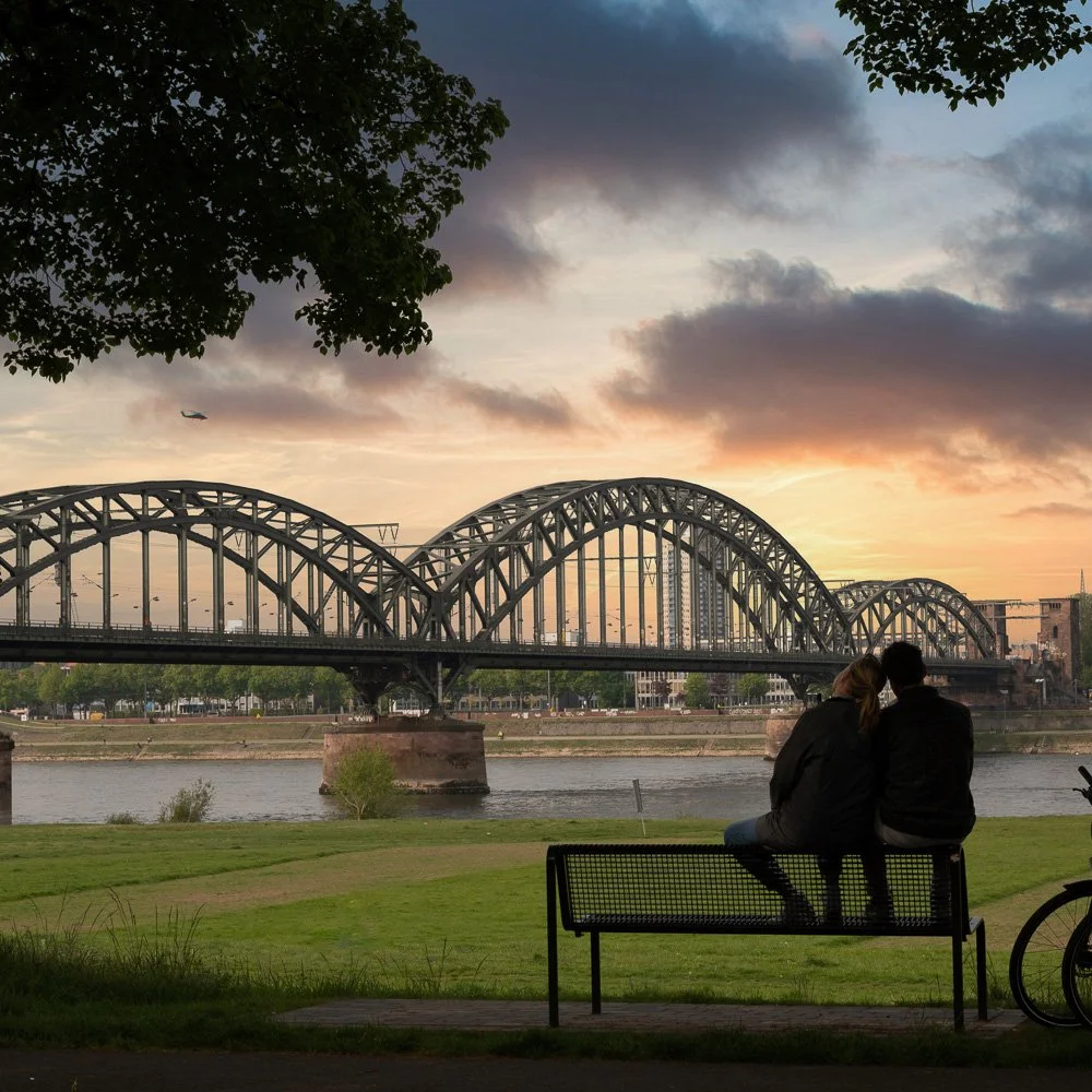 Paar auf einer Bank am Rheinufer bei Sonnenuntergang vor der Kölner Südbrücke, Fahrräder daneben – Tourismusfotografie in Köln