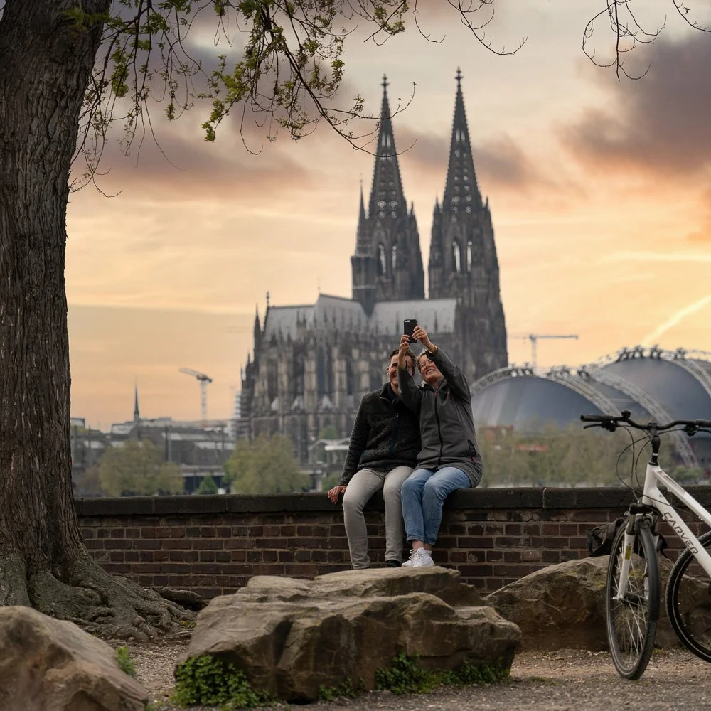 Paar macht ein Selfie auf einer Mauer vor dem Kölner Dom bei Sonnenuntergang, Fahrrad daneben – Reisefotografie in Köln