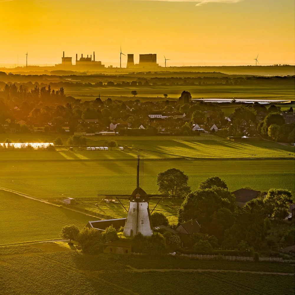 Drohnenaufnahme einer Windmühle im Sonnenaufgang, Dorf und Kraftwerks-Kühltürme als Silhouette am Horizont.