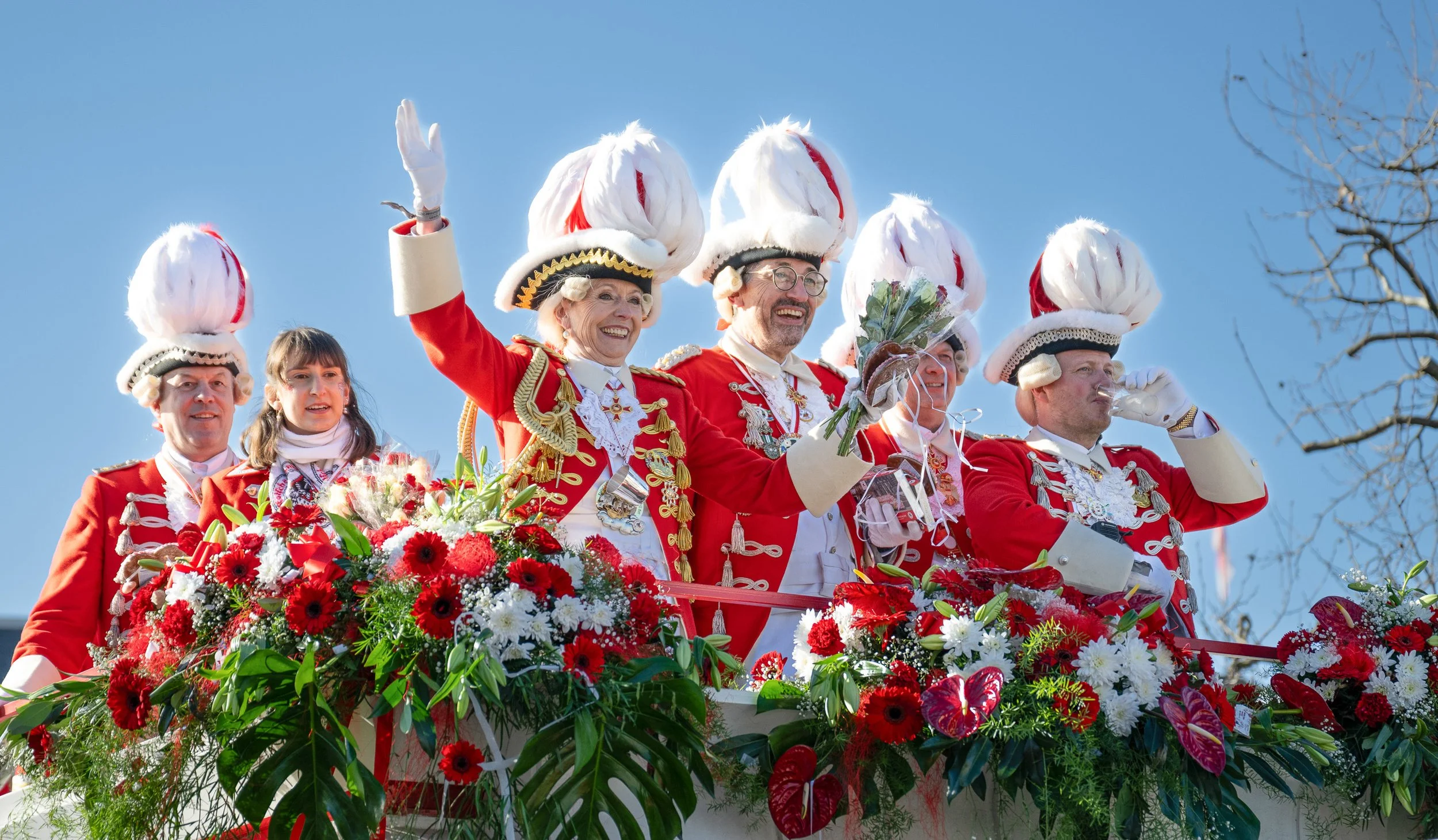 Karnevalsgarde in roter Uniform mit weißen Federhüten unter blauem Himmel.