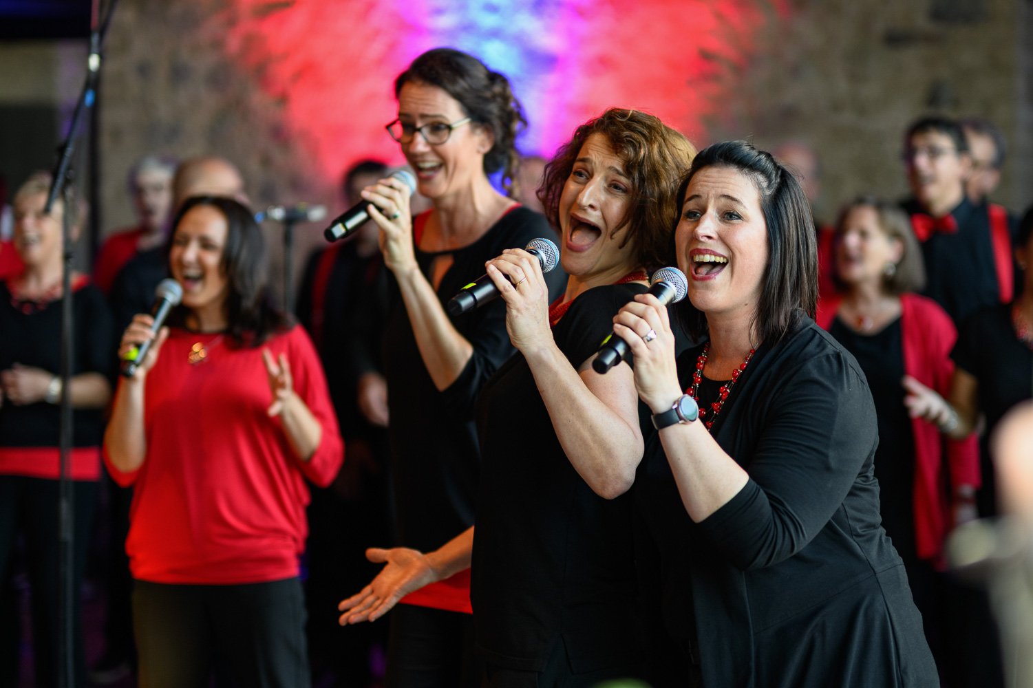 Gesangsensemble singt gemeinsam in Mikrofone im roten Bühnenlicht.