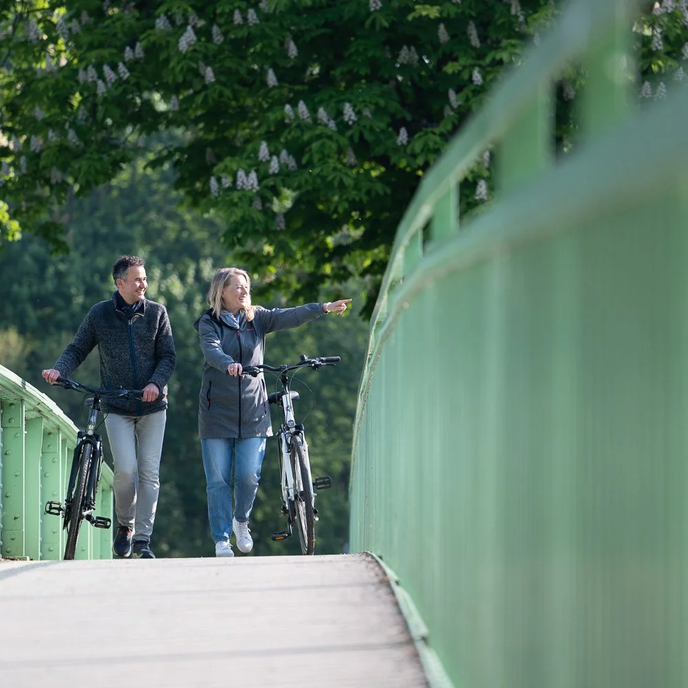 Paar schiebt Fahrräder über eine grüne Bogenbrücke zwischen blühenden Kastanien – Tourismusfotografie im Frühling