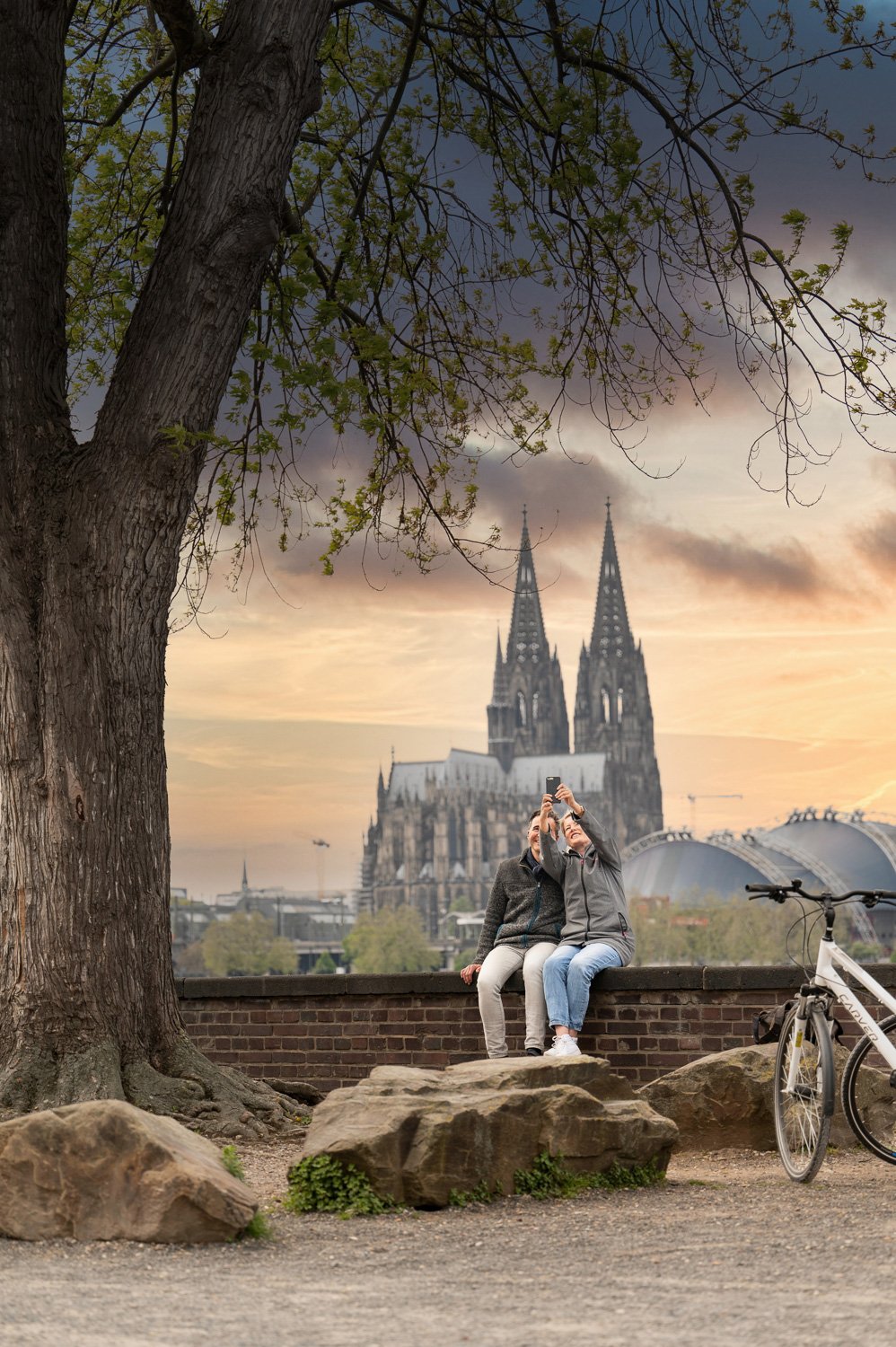 Paar macht ein Selfie auf einer Mauer vor dem Kölner Dom bei Sonnenuntergang, Fahrrad daneben – Reisefotografie in Köln