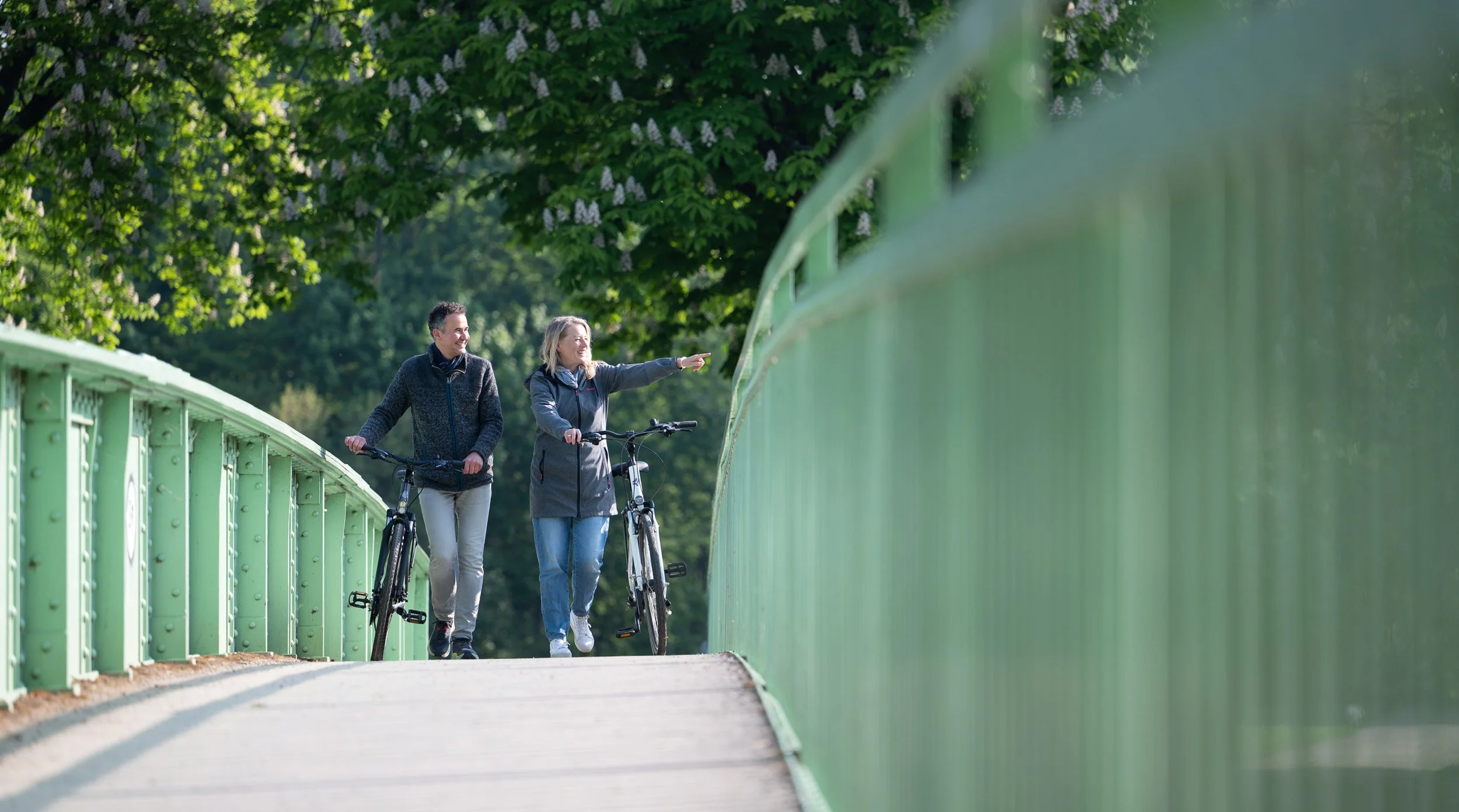 Paar schiebt Fahrräder über eine grüne Bogenbrücke zwischen blühenden Kastanien – Tourismusfotografie im Frühling