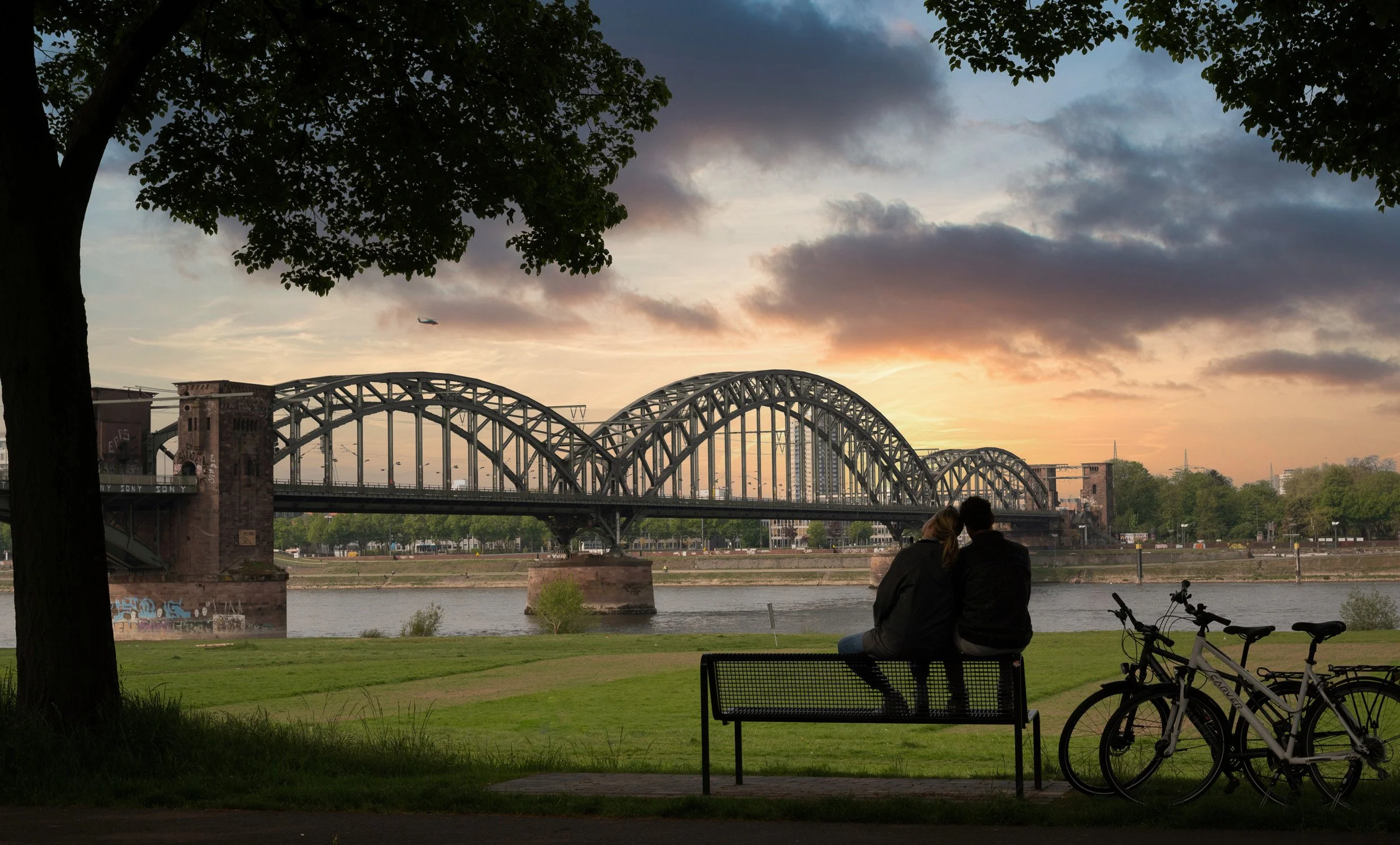 Paar auf einer Bank am Rheinufer bei Sonnenuntergang vor der Kölner Südbrücke, Fahrräder daneben – Tourismusfotografie in Köln