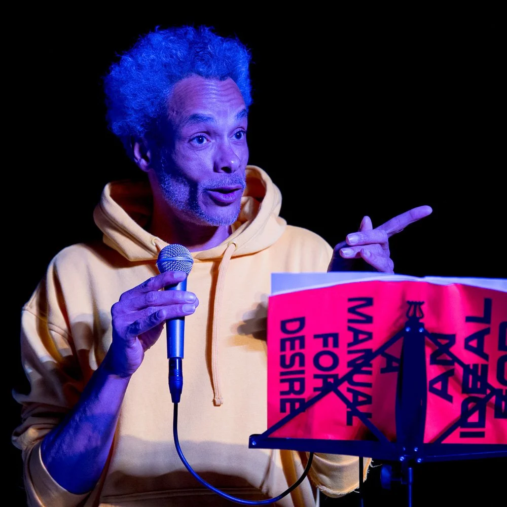 Performance-Künstler am Pult mit Schild in blauem Bühnenlicht.