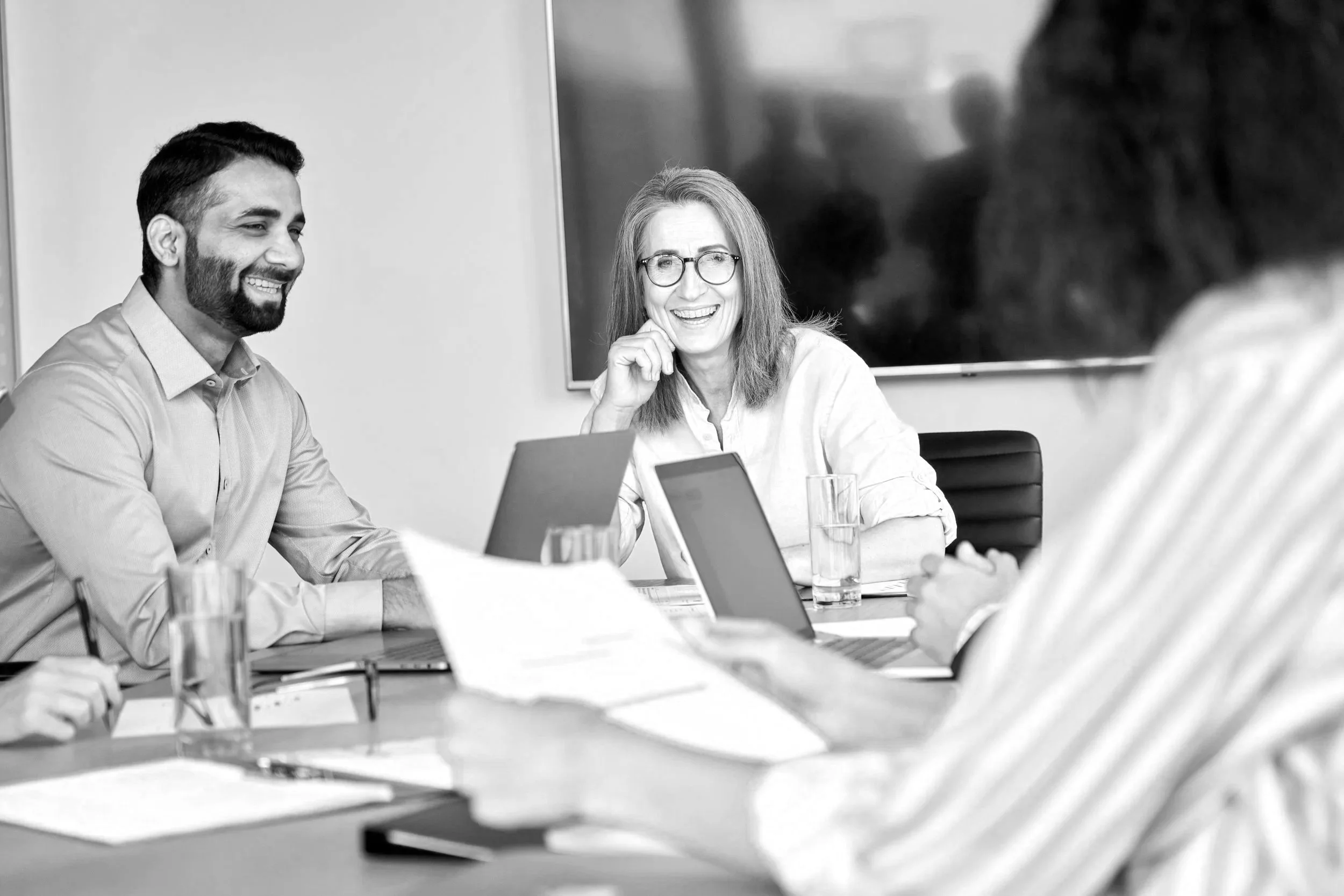 Group of people in a meeting room, smiling and engaging in conversation, with laptops and documents on the table.