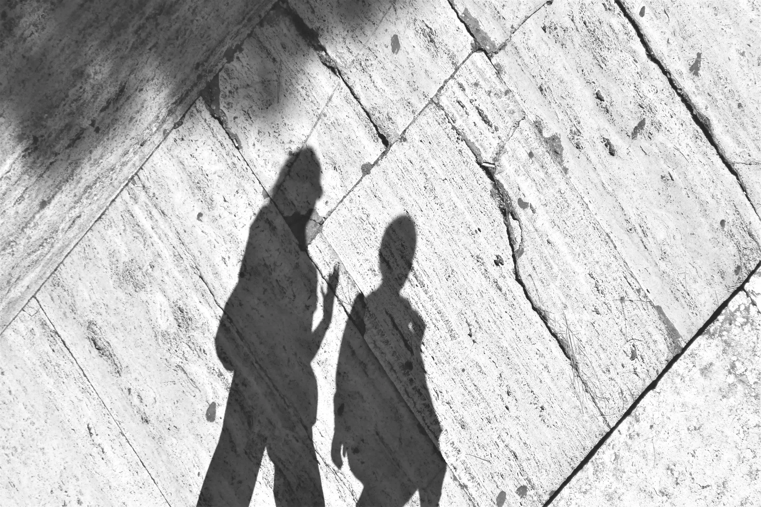 Shadows of two people walking on a stone sidewalk, with visible cracks and textured surface, cast by sunlight.