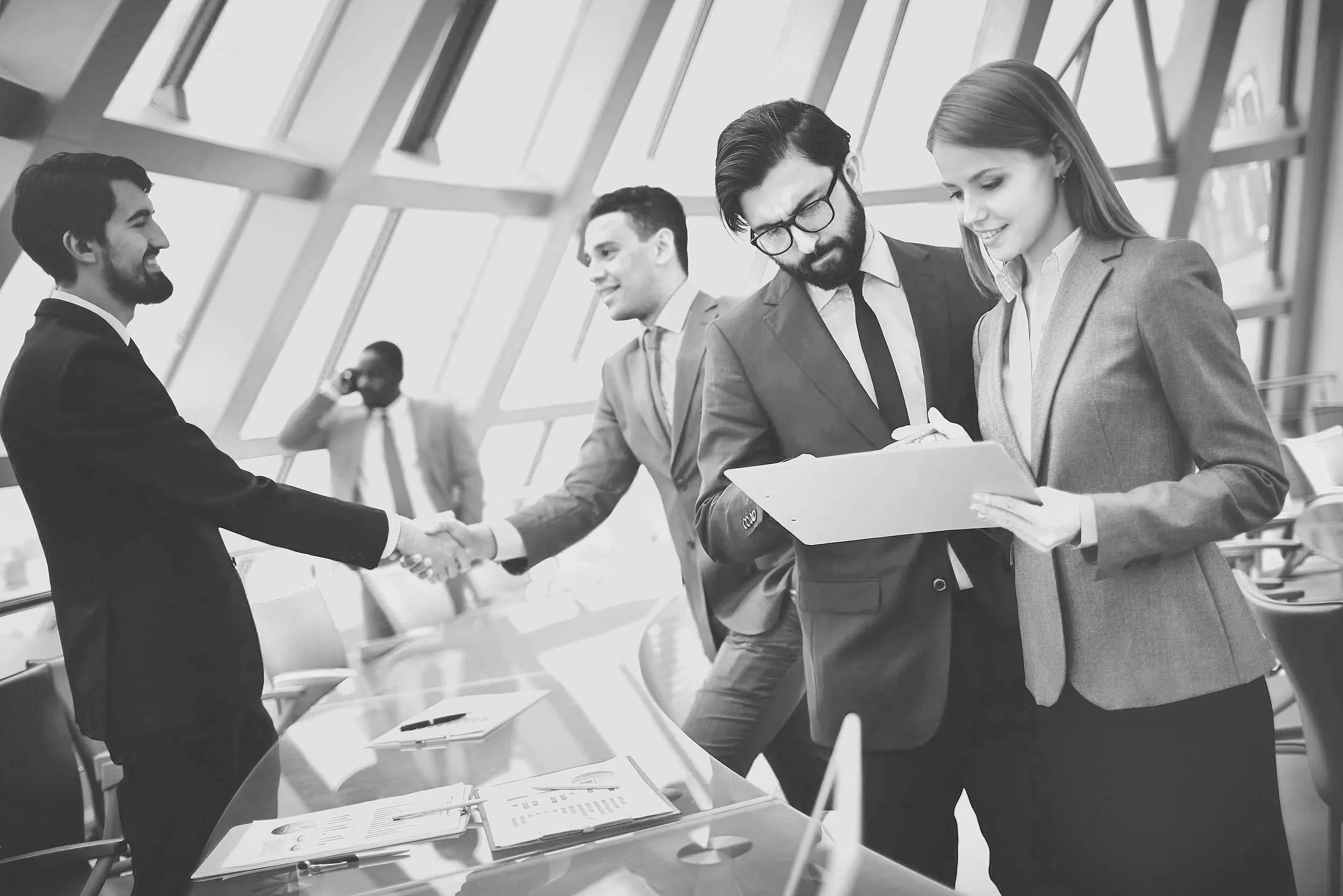 Business professionals in a modern office engaging in a handshake and reviewing documents, with a person in the background on the phone, all dressed in formal attire.