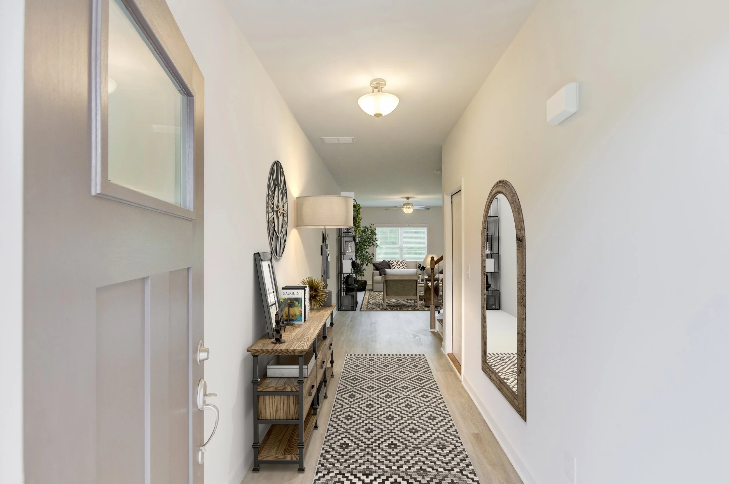 A hallway in a home with light-colored walls and hardwood flooring. It features a long, patterned rug, a wooden console table with decorative items, a large wall clock, a mirror, and a tall lamp. The hallway leads to a living room with large windows, a ceiling fan, and furniture including sofas and a ceiling fan.