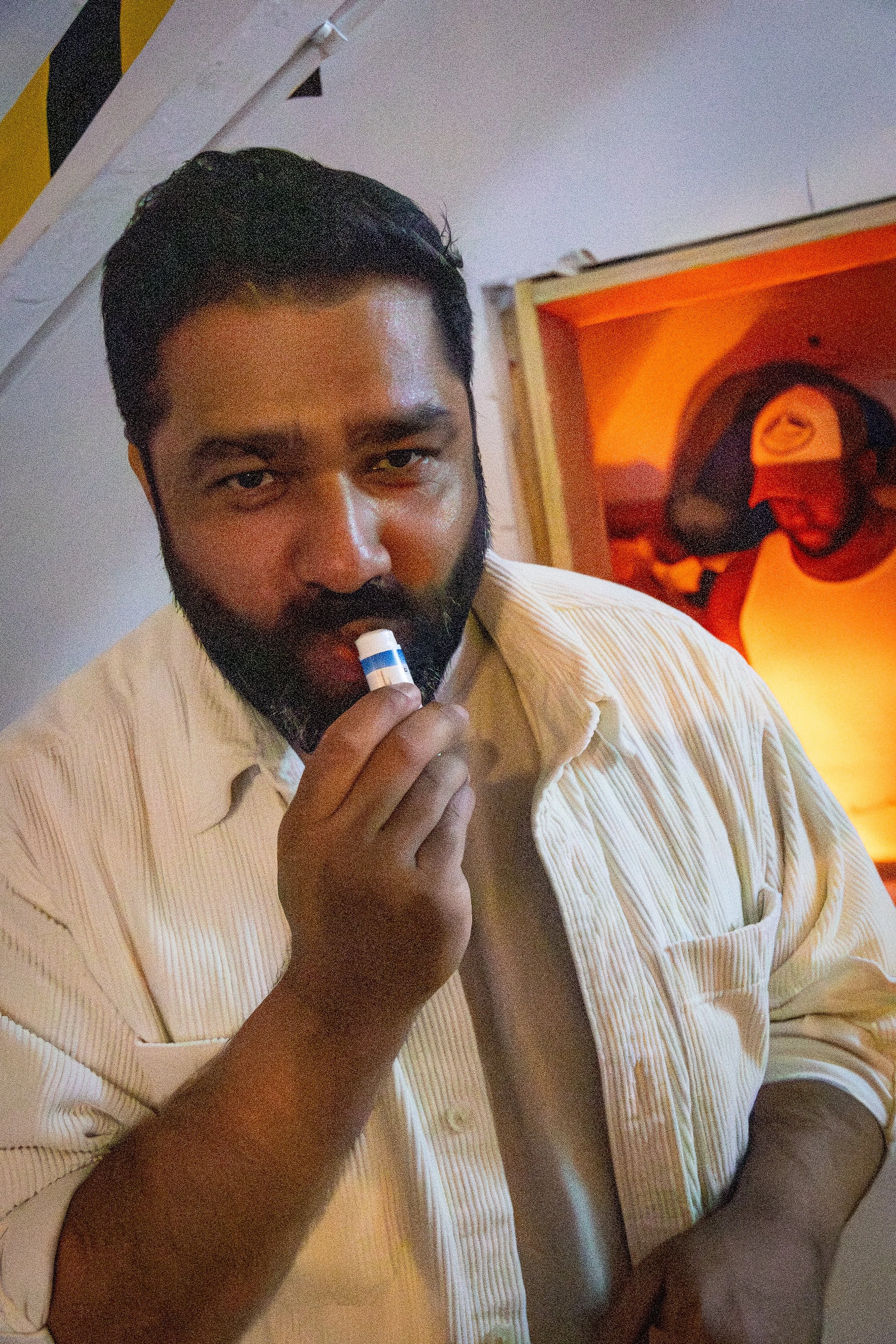 A man with dark hair and a beard in a light-colored, vertically striped shirt is holding an inhaler near his mouth. Behind him, there is a partially visible painting of two men, one of whom is wearing sunglasses and a cap, with an orange background. 