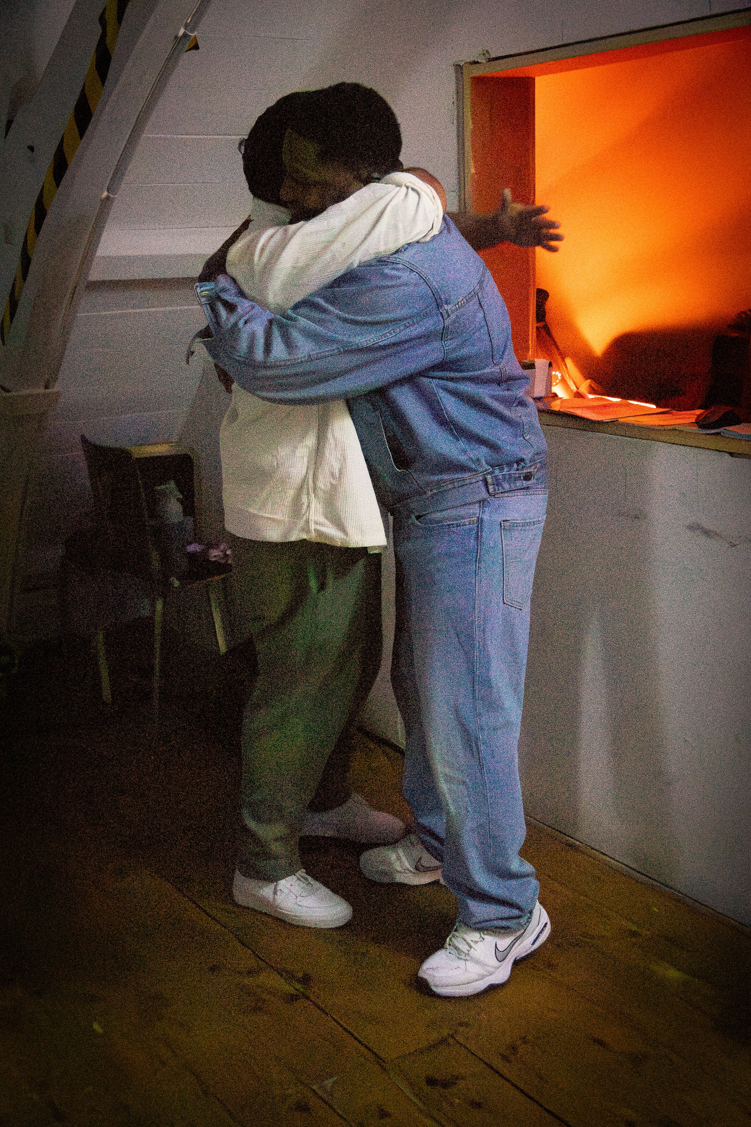 Two people embracing each other in a hug inside a room with orange and white walls. They are standing on a wooden floor, wearing casual clothes and white sneakers.