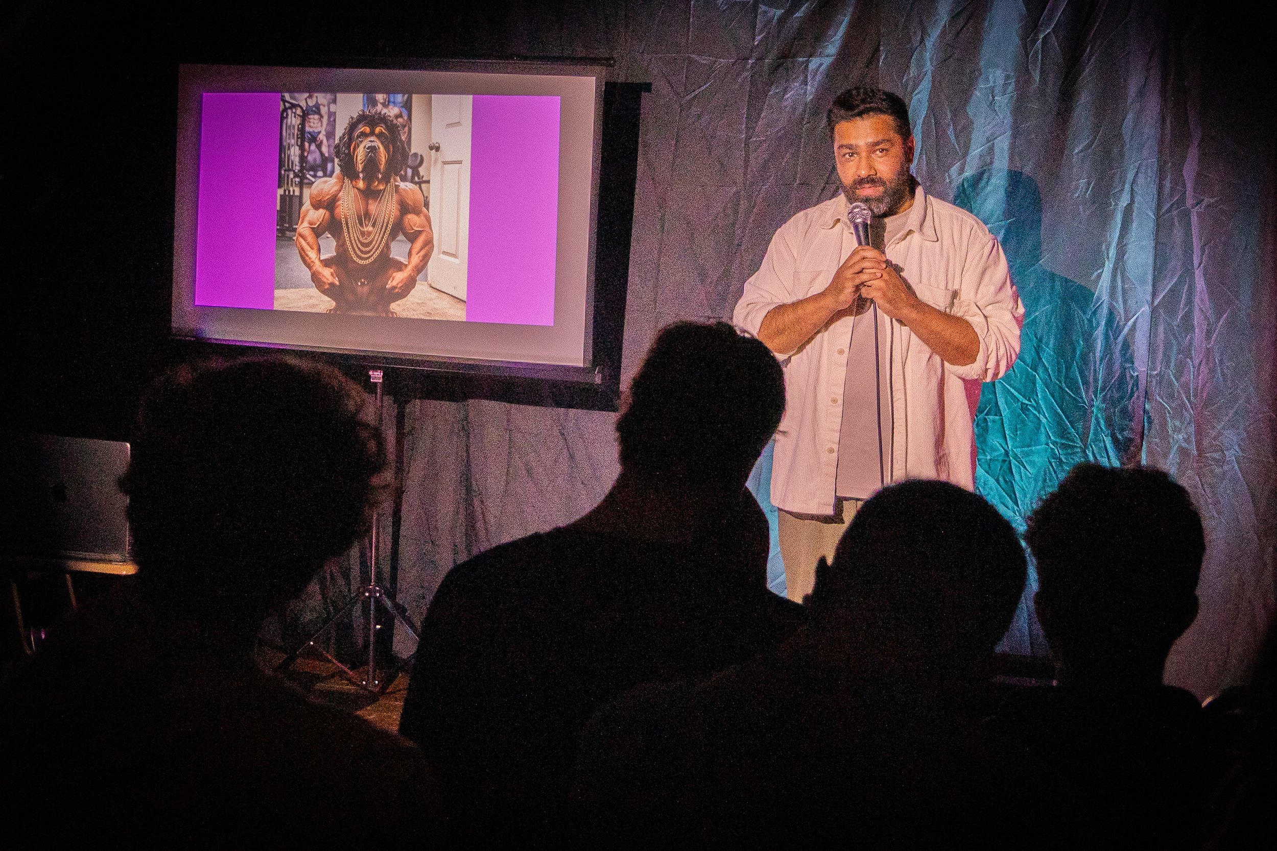 A man with a beard holding a microphone, standing in front of a projection screen. The screen displays a humorous photo of a dog with a human body, wearing jewelry, in a gym setting.