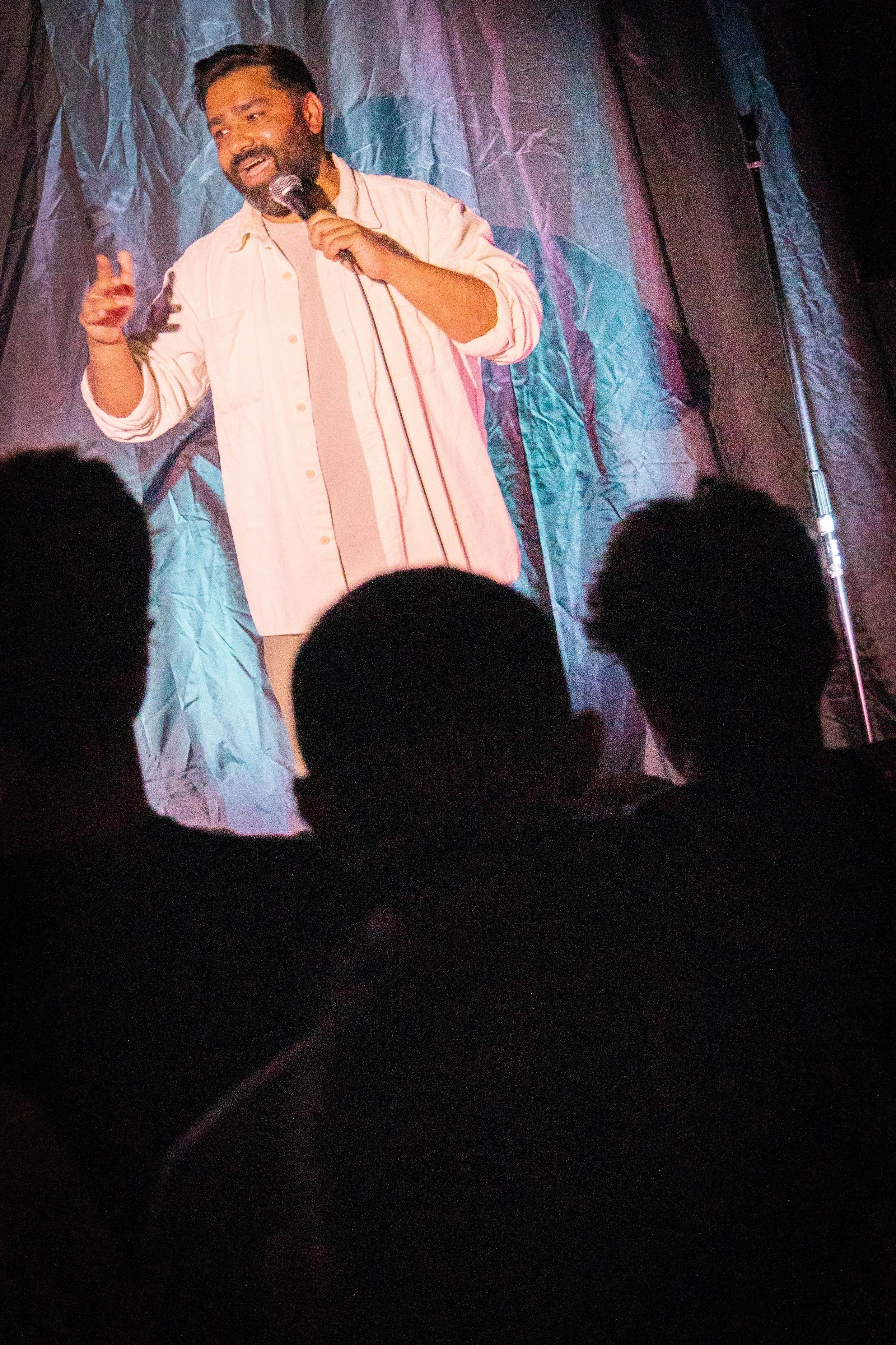 Comedian standing on stage, holding microphone, wearing light-colored shirt, speaking to audience in a dimly lit venue.