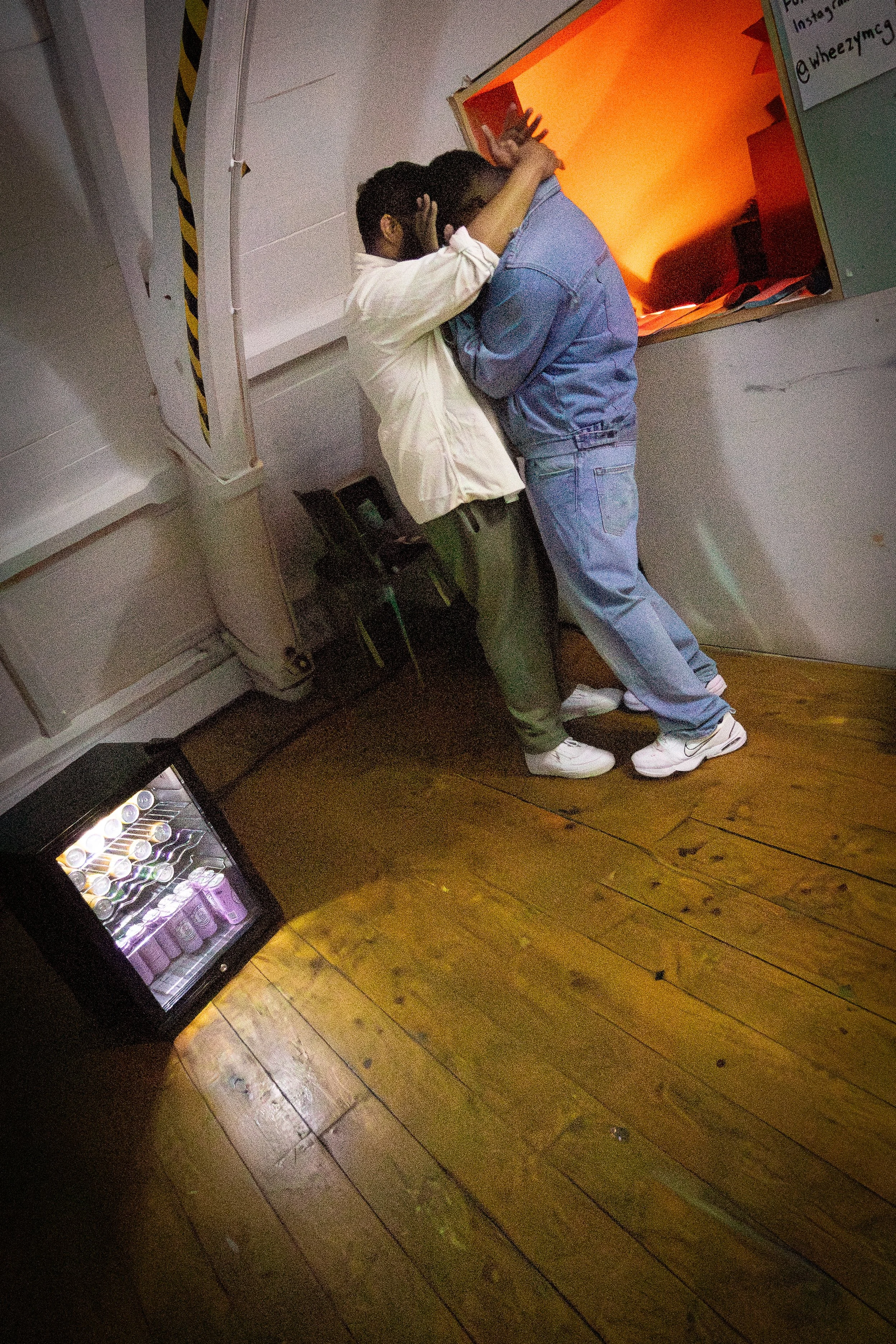 Two boys are standing close together, with one leaning their head on the other's shoulder, in front of an orange-lit mirror or window. They appear to be hugging or comforting each other.