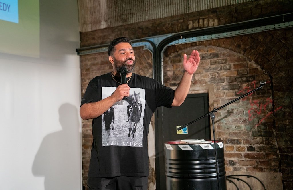 A man with a beard and short hair speaking into a microphone, gesturing with one hand, in front of a brick wall and a large screen.