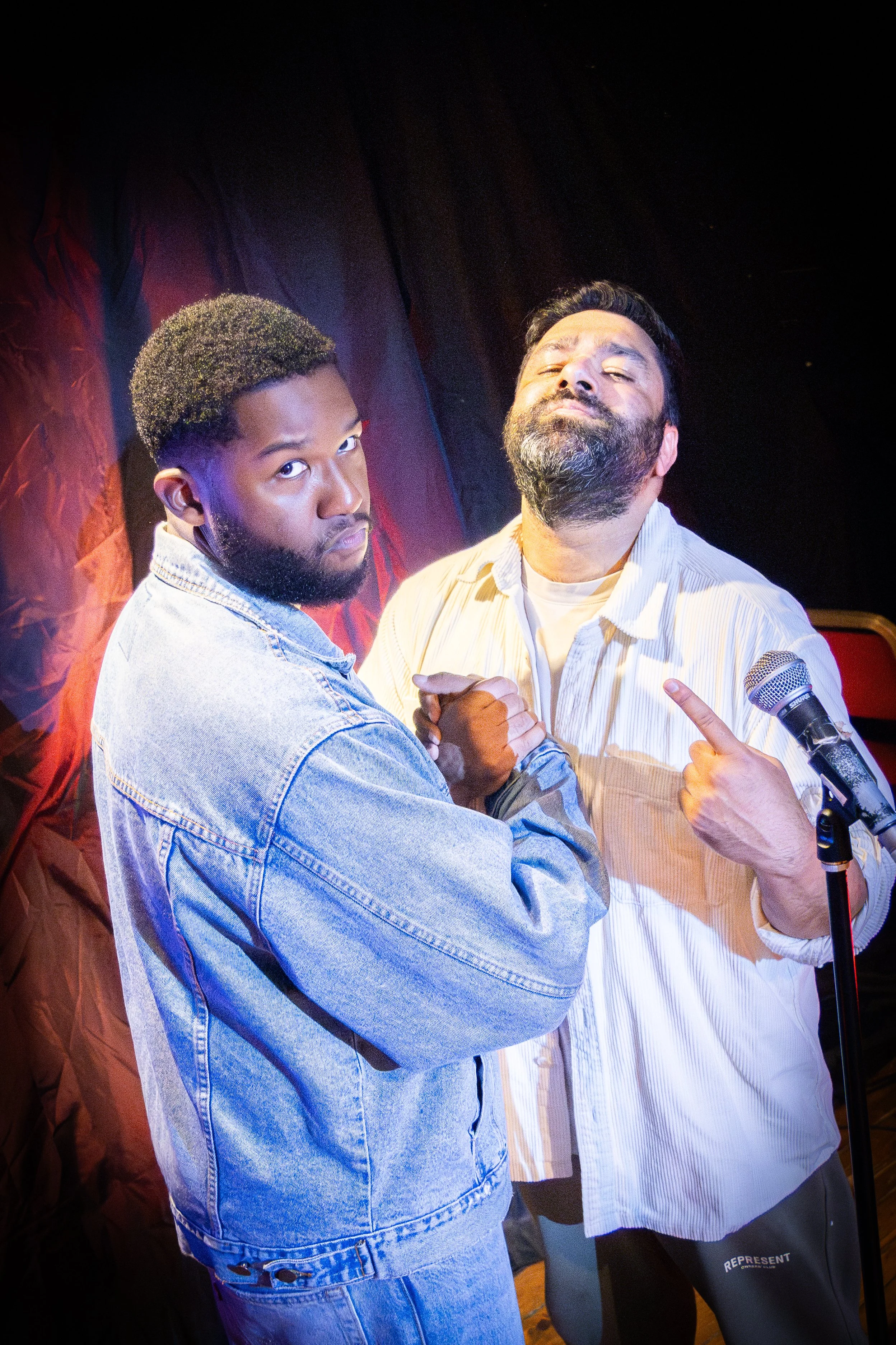 Two men facing each other arm wrestling, with one man wearing a denim jacket and the other in a shirt, in a performance or event setting with a microphone on a stand nearby.