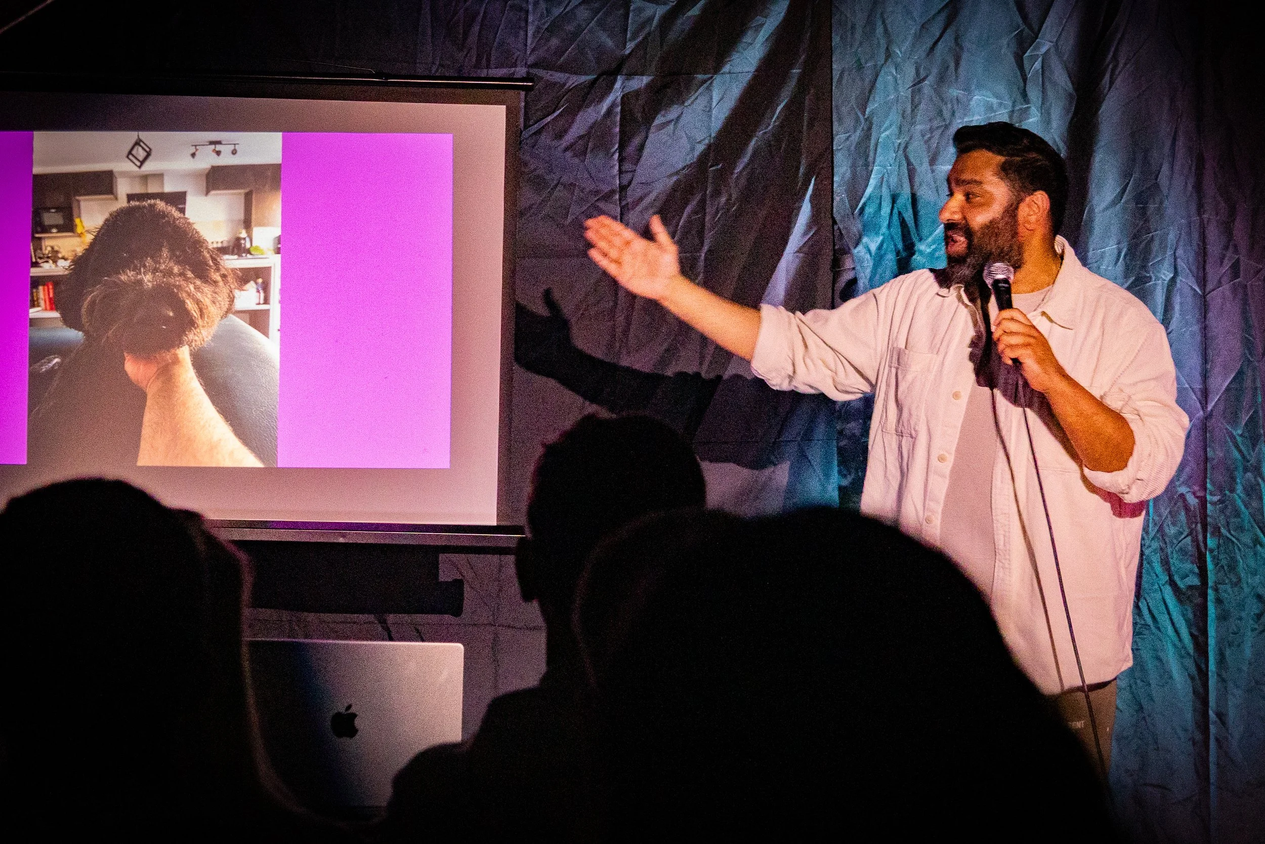 A man holding a microphone and gesturing towards a presentation screen with a photo of a person with curly hair, in a room with dark textured walls and an audience.