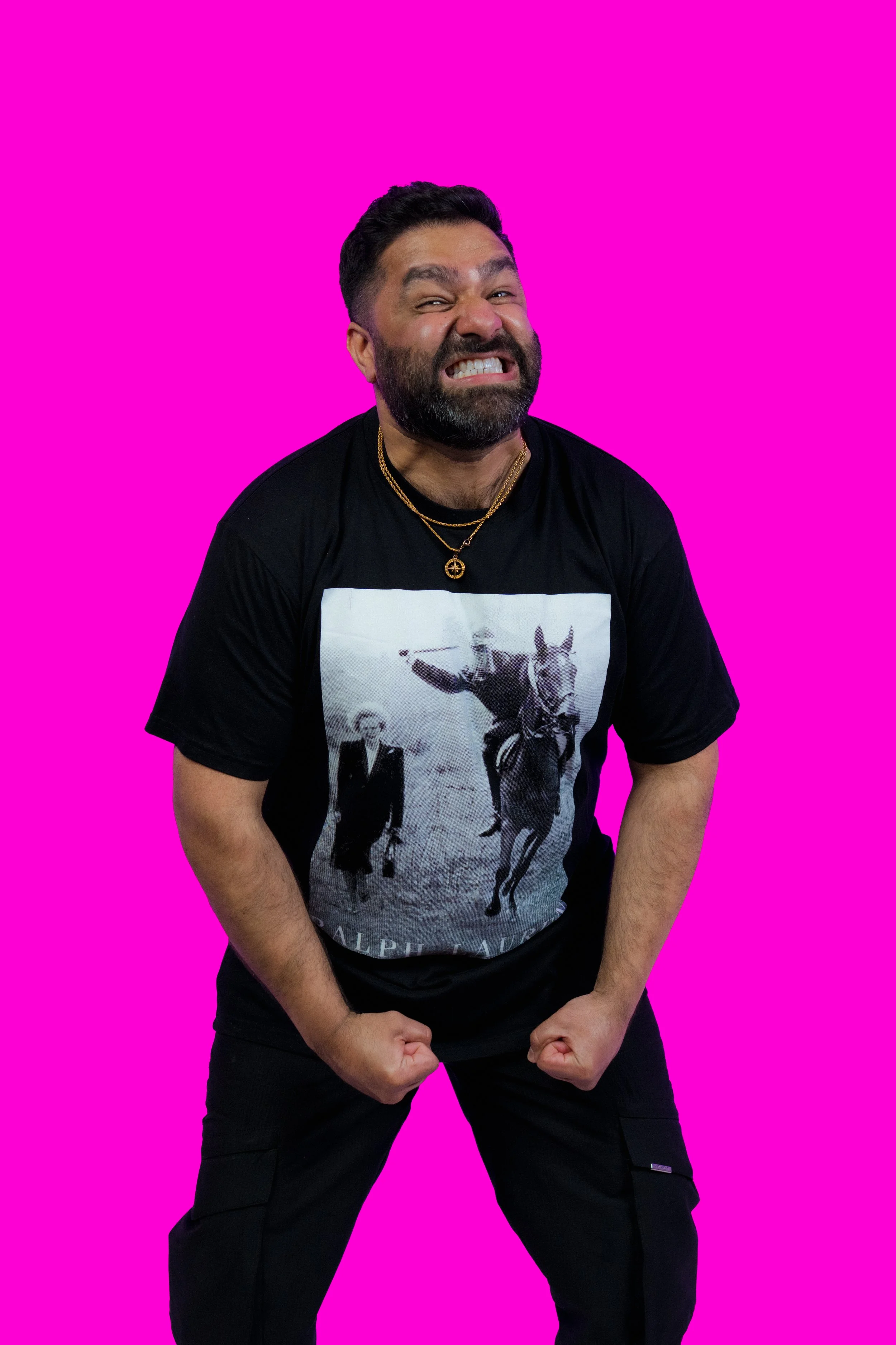 A man with dark hair and a beard making a muscular pose, wearing a graphic black t-shirt and gold necklaces, standing in front of a pink background.