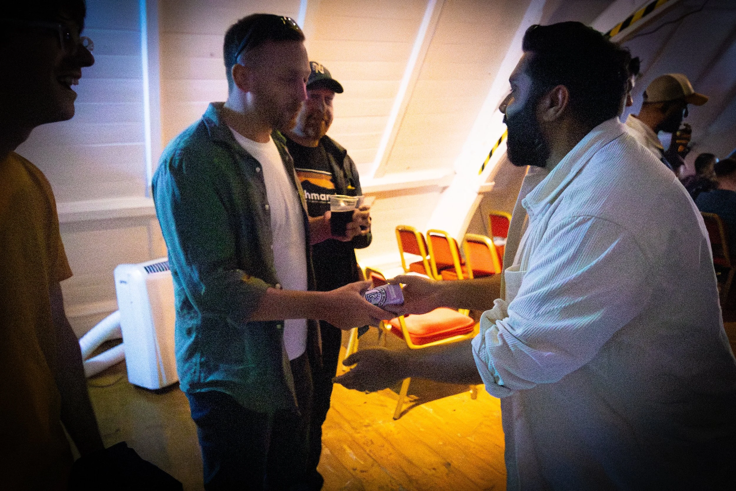 Two men shaking hands, one wearing a white shirt and the other in a dark green jacket with a white shirt beneath. They are indoors with yellow chairs and a white ceiling in the background, and there are other people around.