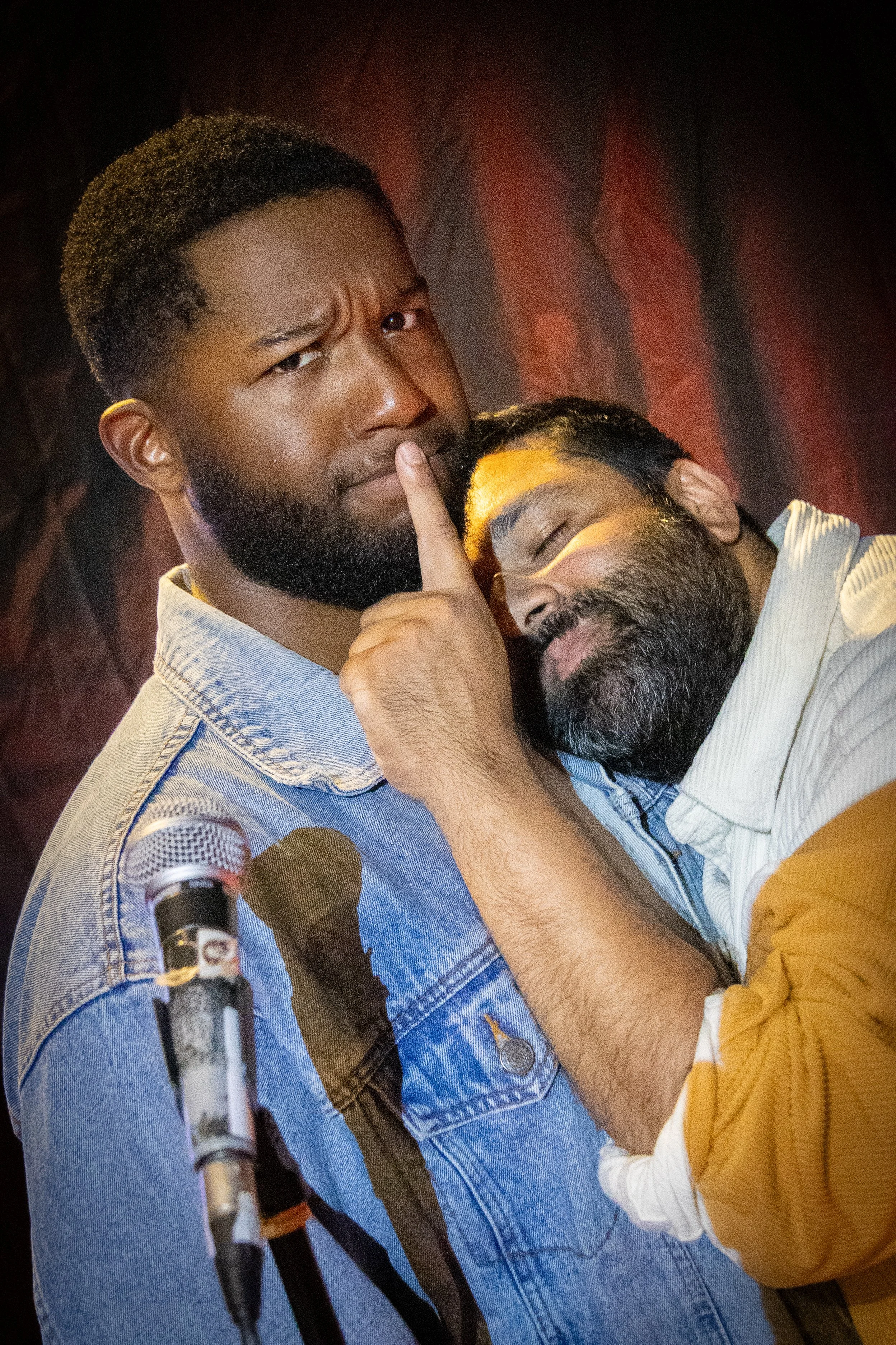 Two men are in a close, affectionate pose. The man on the right has his hand on the other's chin and appears playful. The background is dark with a reddish hue.