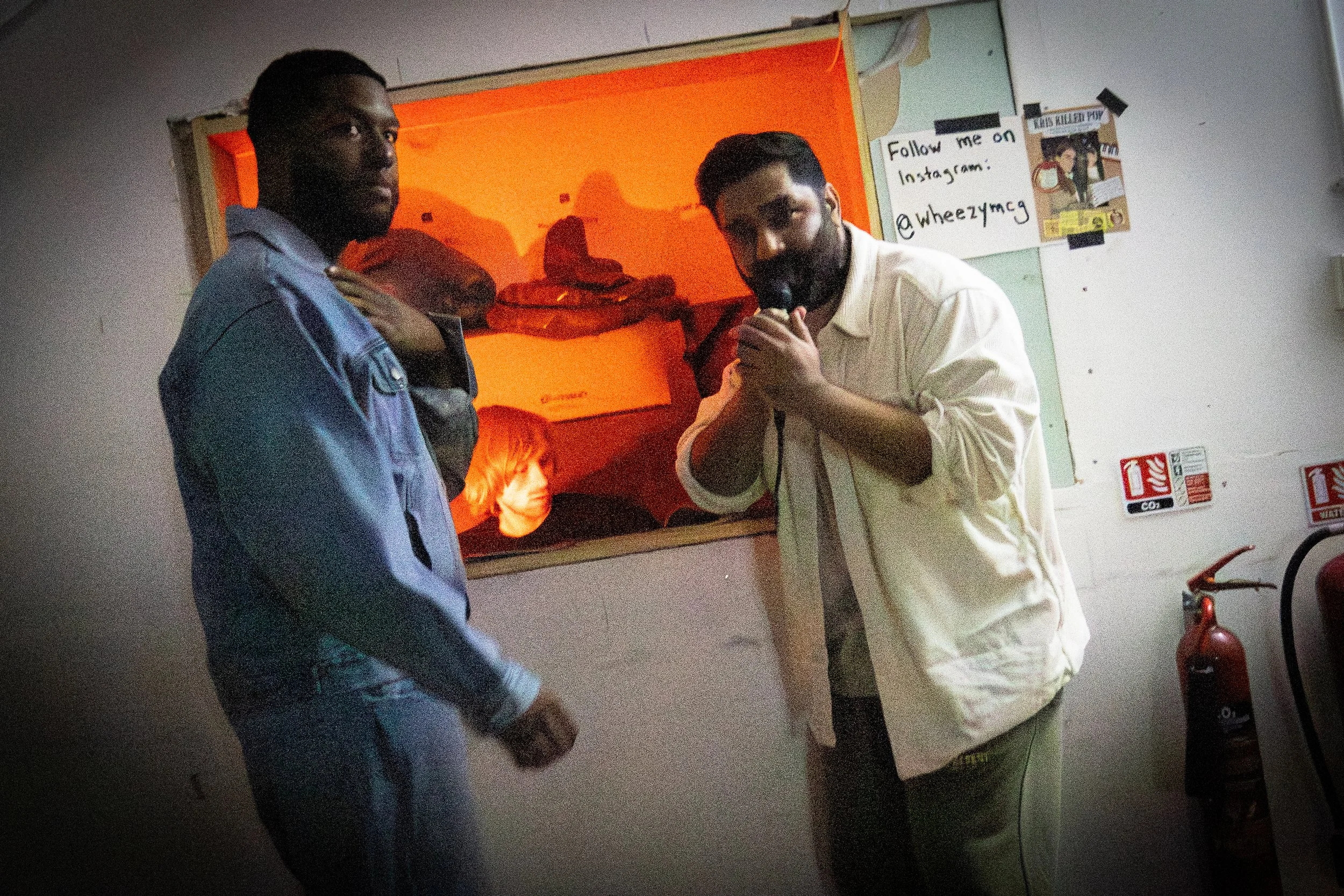 Two men, one with a microphone, inside a room with a marked wall and fire extinguishers.