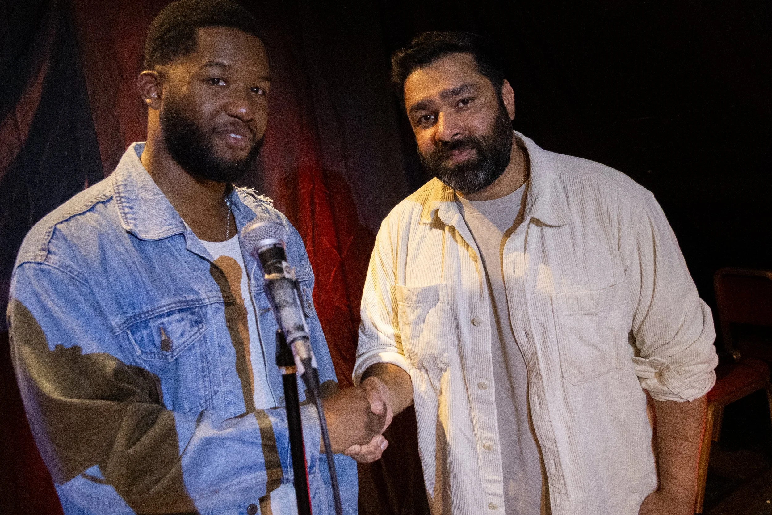 Two men shaking hands, one is holding a microphone. They are standing in a dark-lit room with black and red curtains in the background.