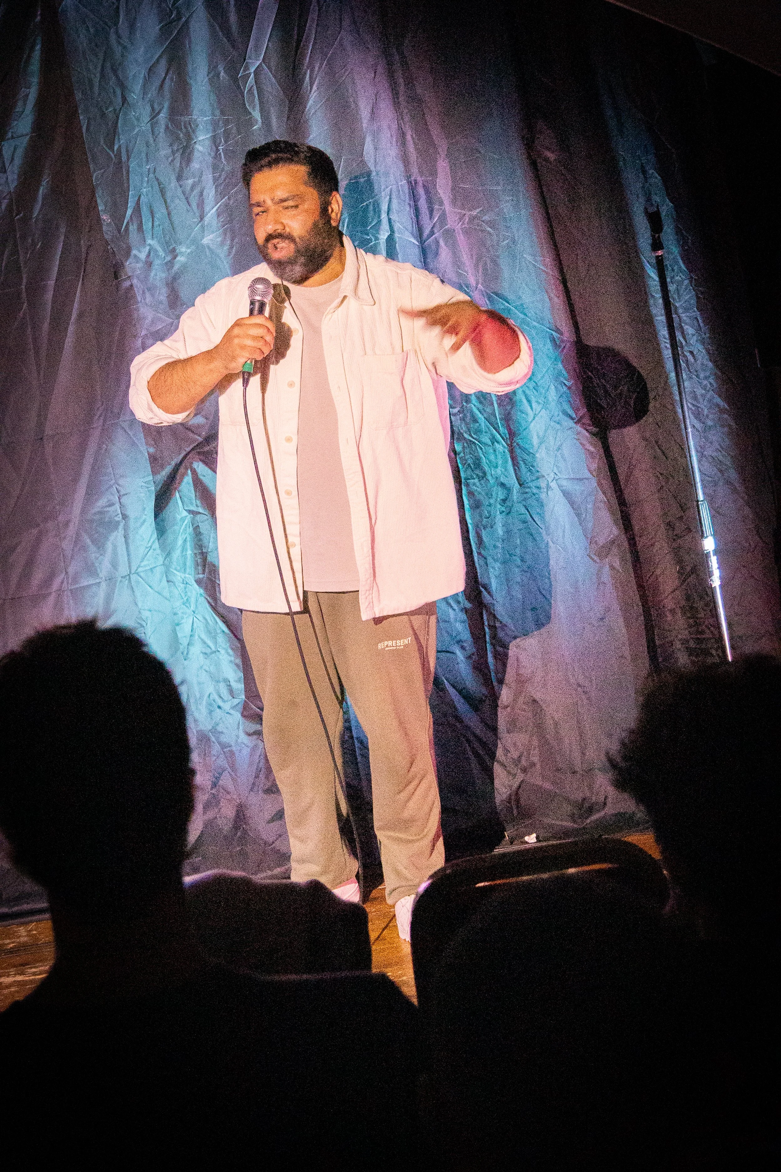 A man with a beard and dark hair performing stand-up comedy or giving a speech on stage, holding a microphone in his right hand, standing in front of a dark textured curtain, with an audience in front.