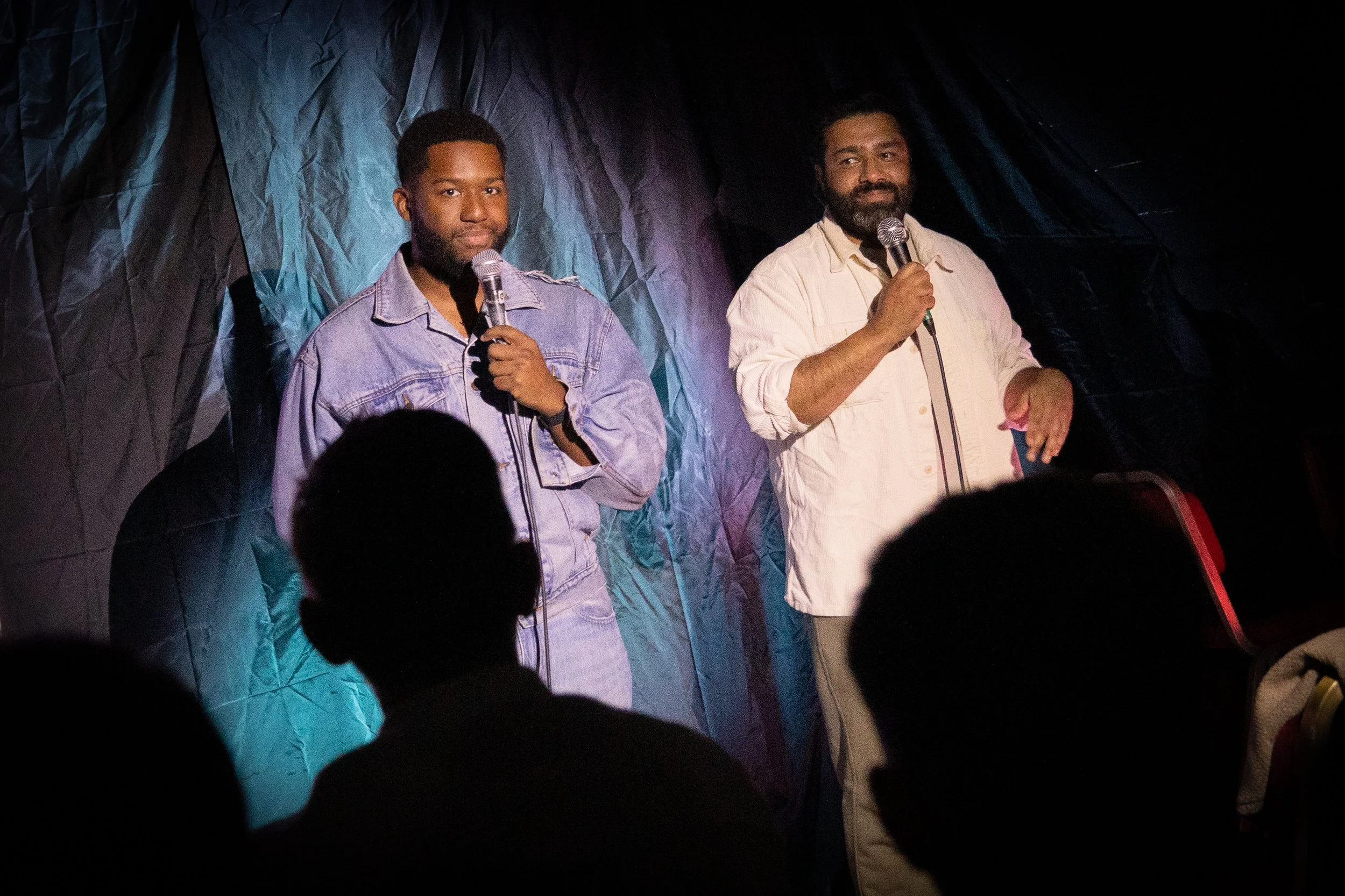 Two men stand on a stage holding microphones, with a dark, crinkled backdrop behind them. The audience is partially visible in the foreground.