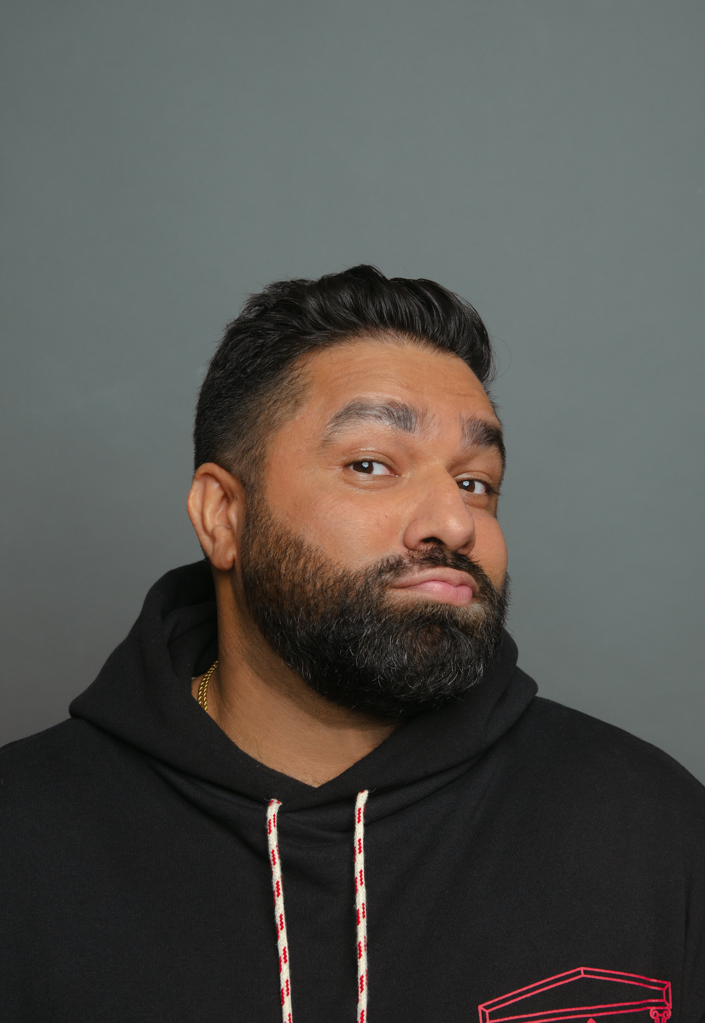 Close-up of a man with dark hair and beard, wearing a black hoodie with red and white drawstrings, against a plain gray background.