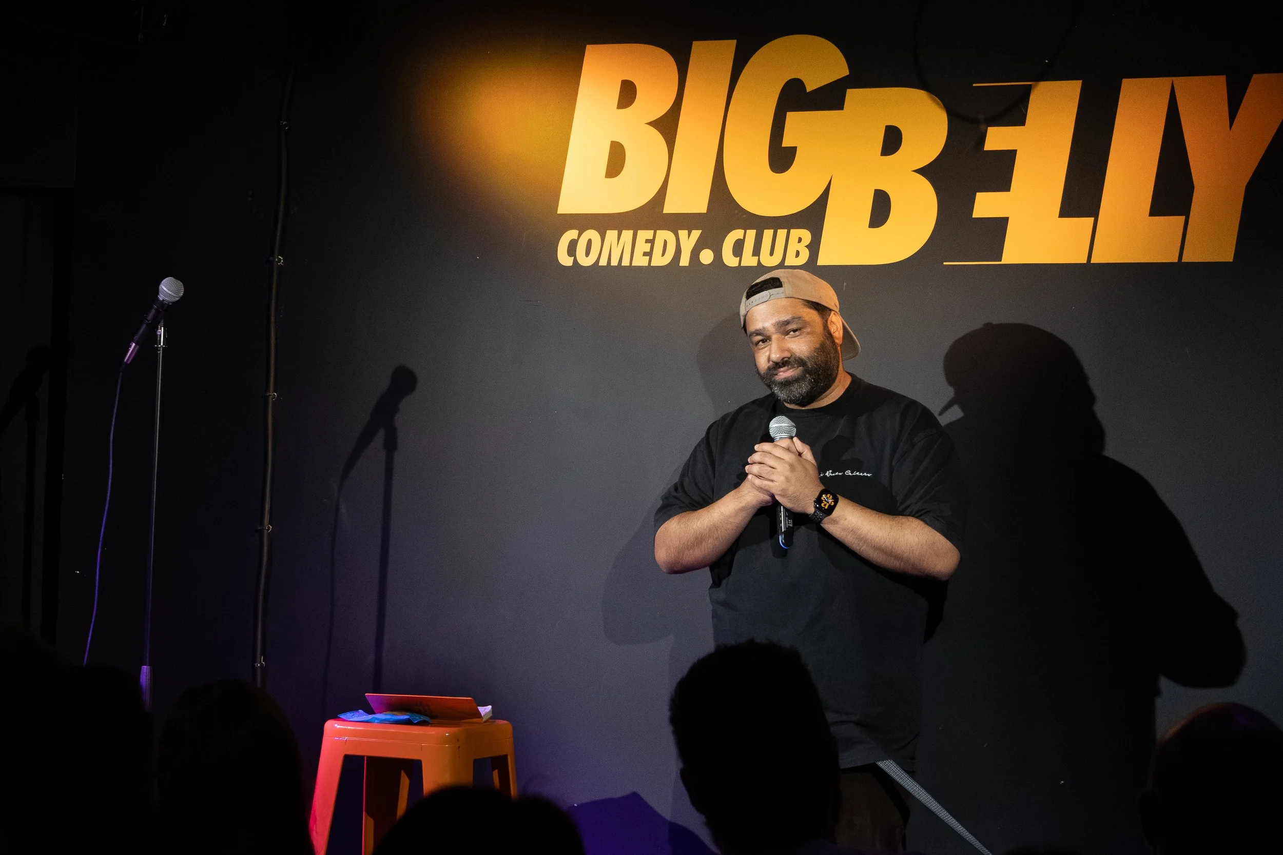 A man stands on stage at Big Belly Comedy Club, holding a microphone, with a black background and large yellow sign displaying the club's name behind him.