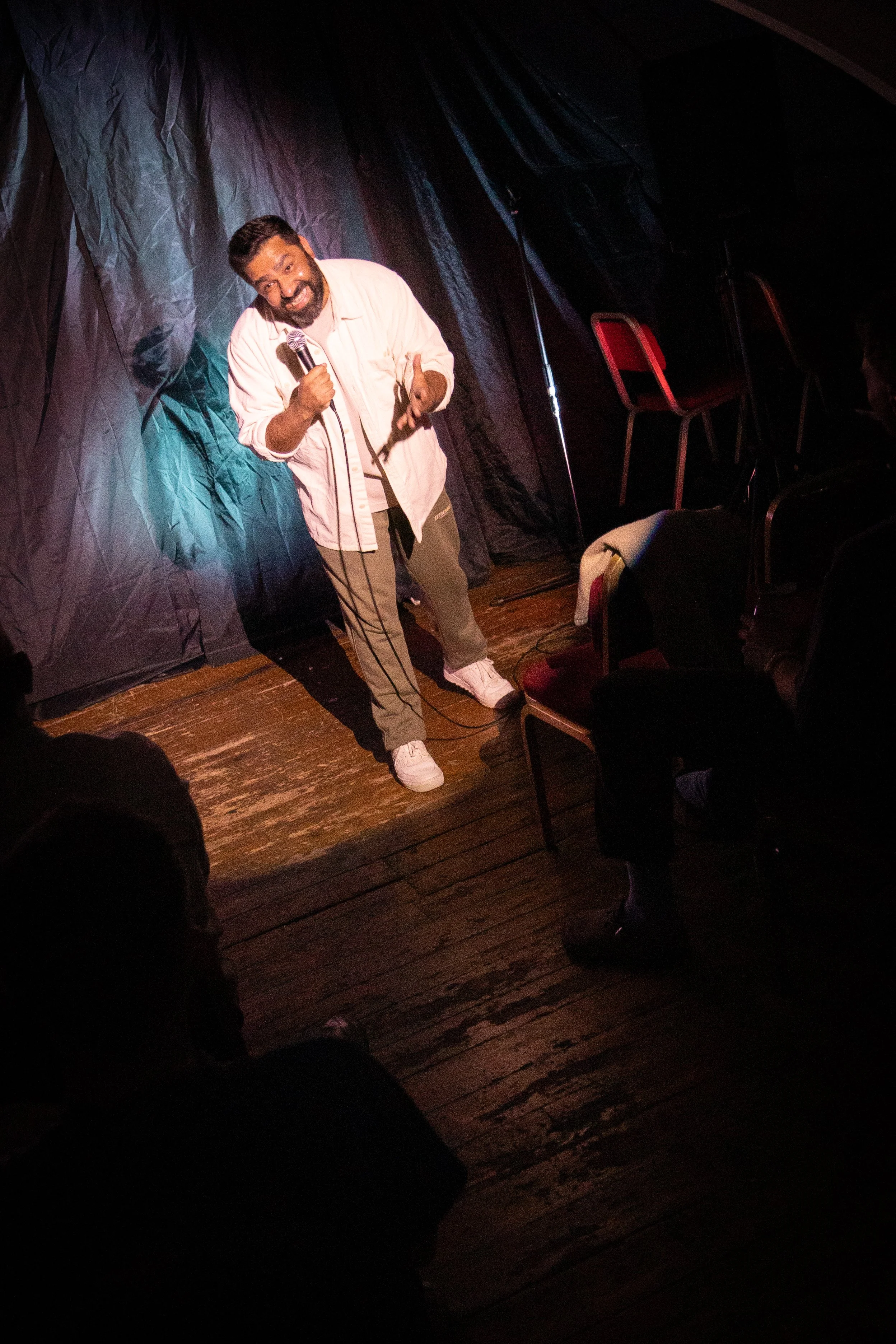 A man with a beard and dark hair is performing stand-up comedy on stage, holding a microphone and smiling. The stage has a dark backdrop and wooden flooring, with some chairs visible to the side.