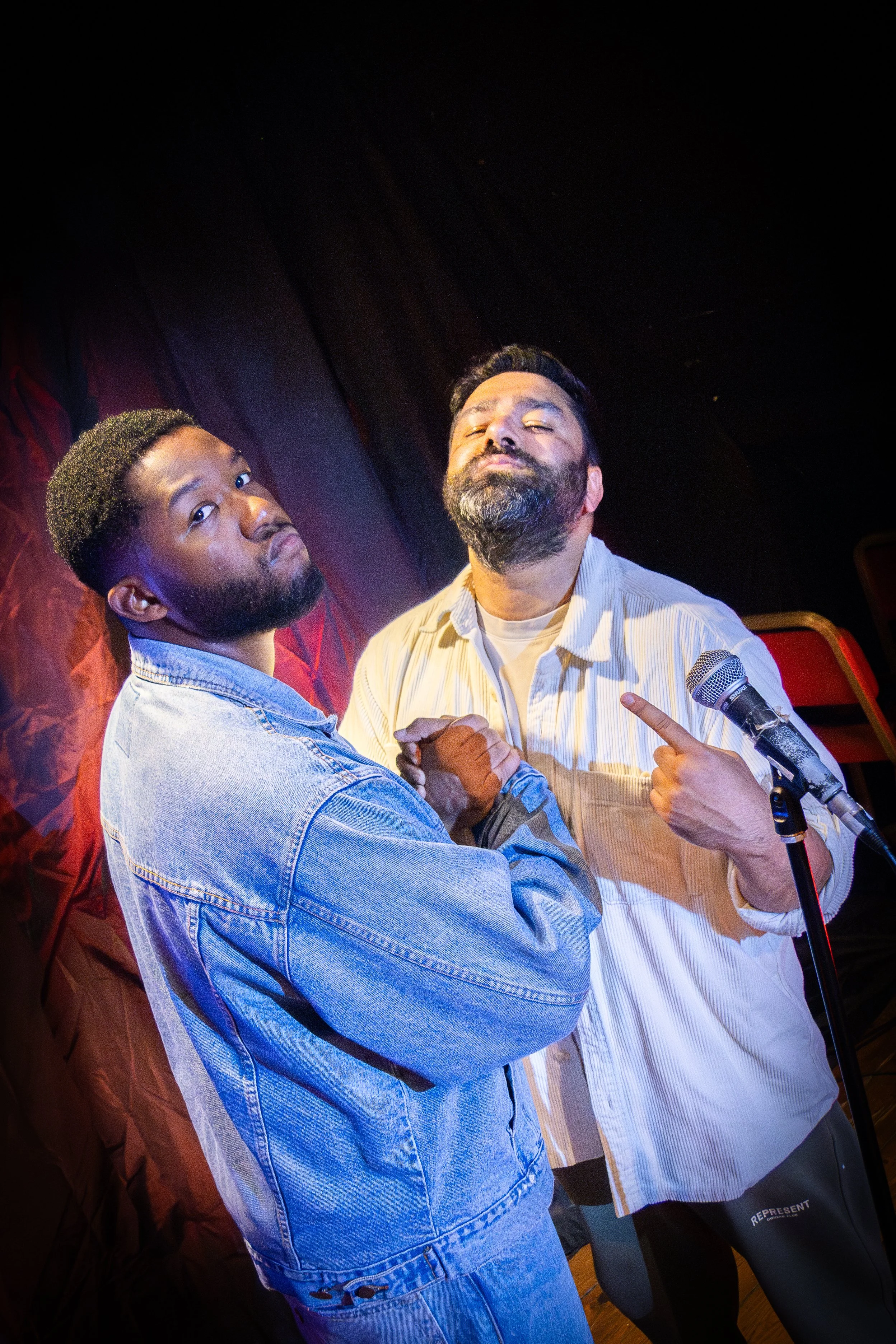 Two men are posing with a confident attitude, one with his arms crossed and the other pointing at himself with a mic nearby. They are in a dimly lit indoor setting, with dark and red background curtains.