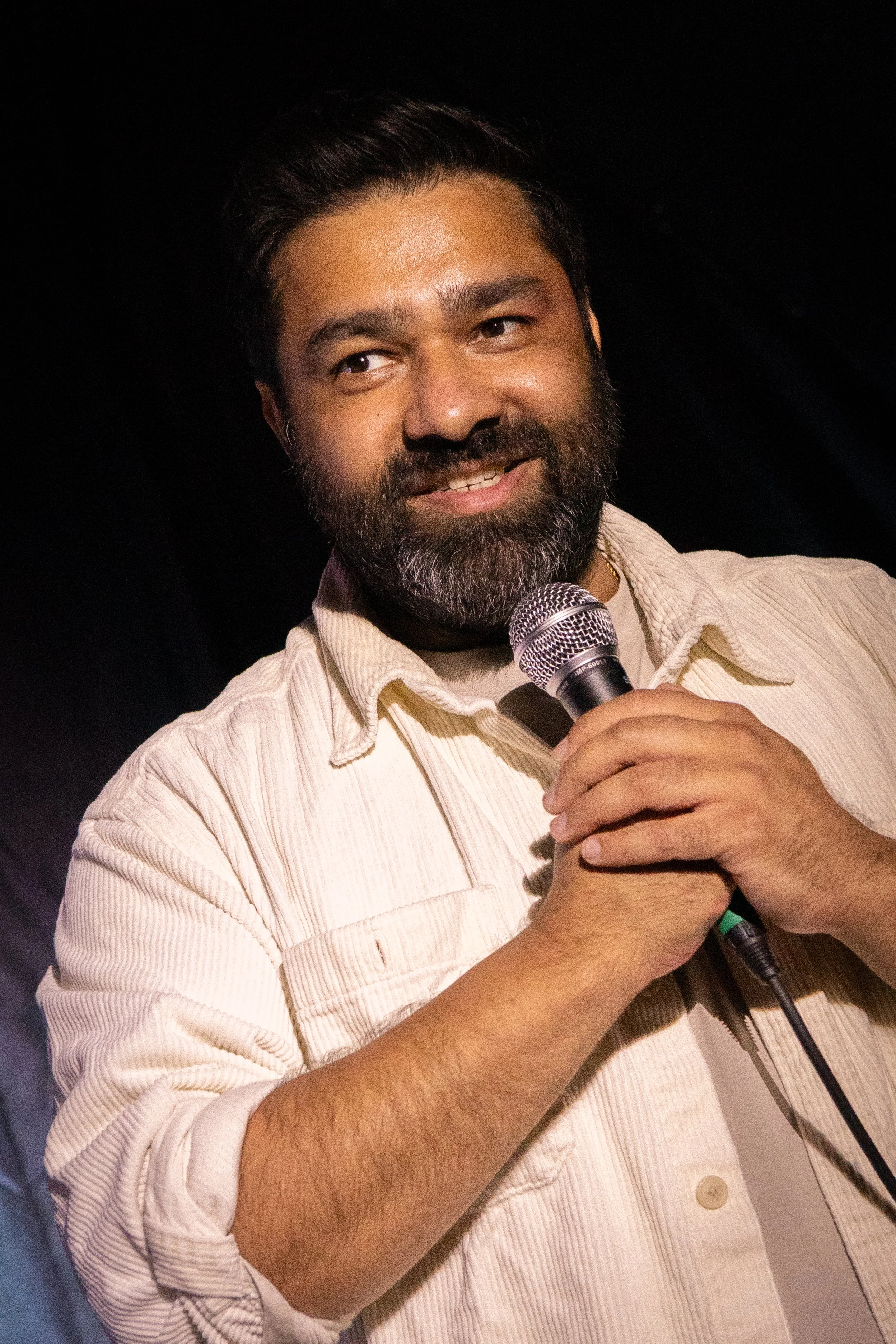 A man with dark hair, a beard, and tan skin holding a microphone and smiling. He is wearing a light-colored, striped button-up shirt.