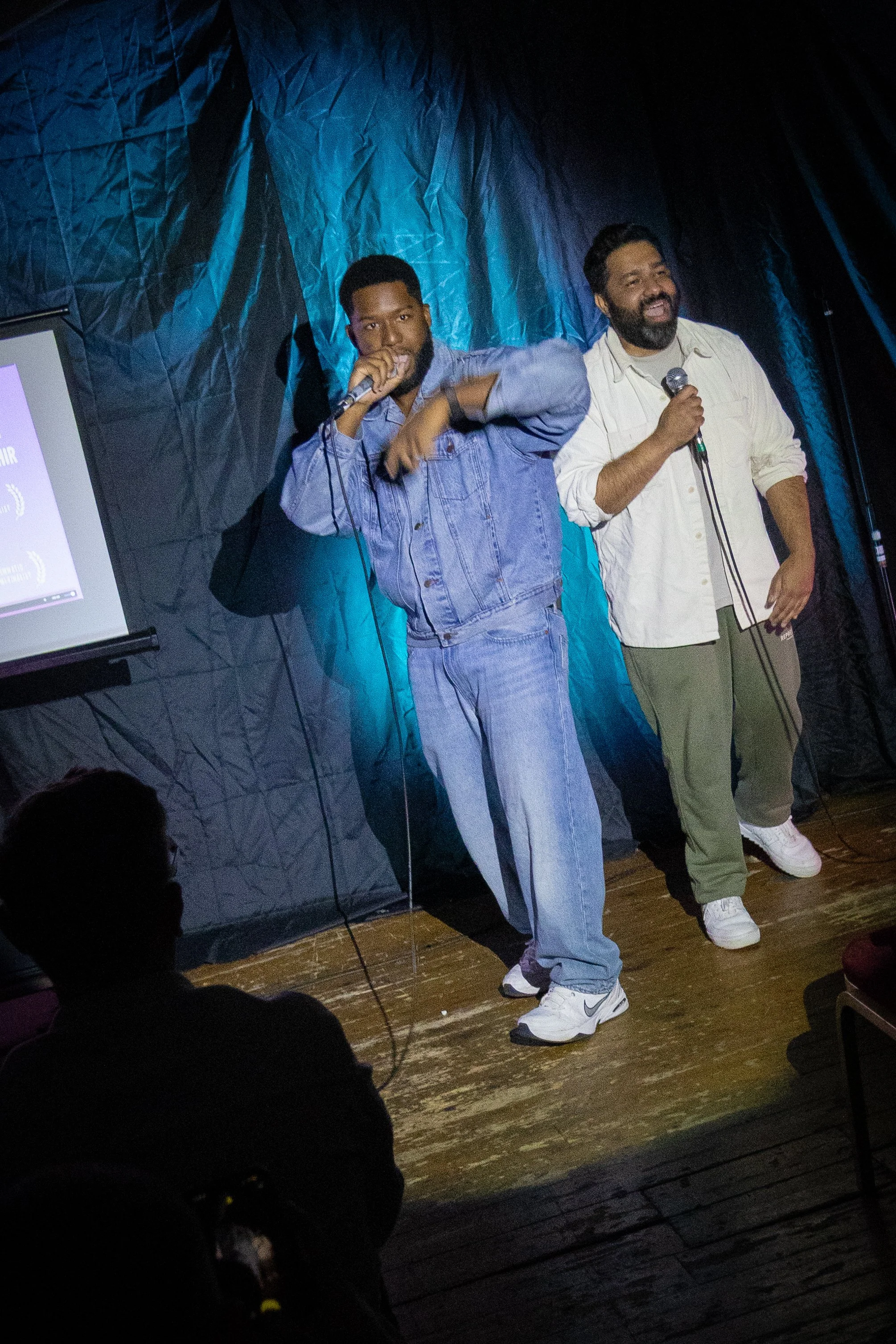 Two men are standing on a wooden stage, holding microphones, engaging with an audience. One is dressed in a denim jacket and jeans, the other in a beige shirt and olive pants. They are performing or speaking, with a dark, gathered backdrop and a part