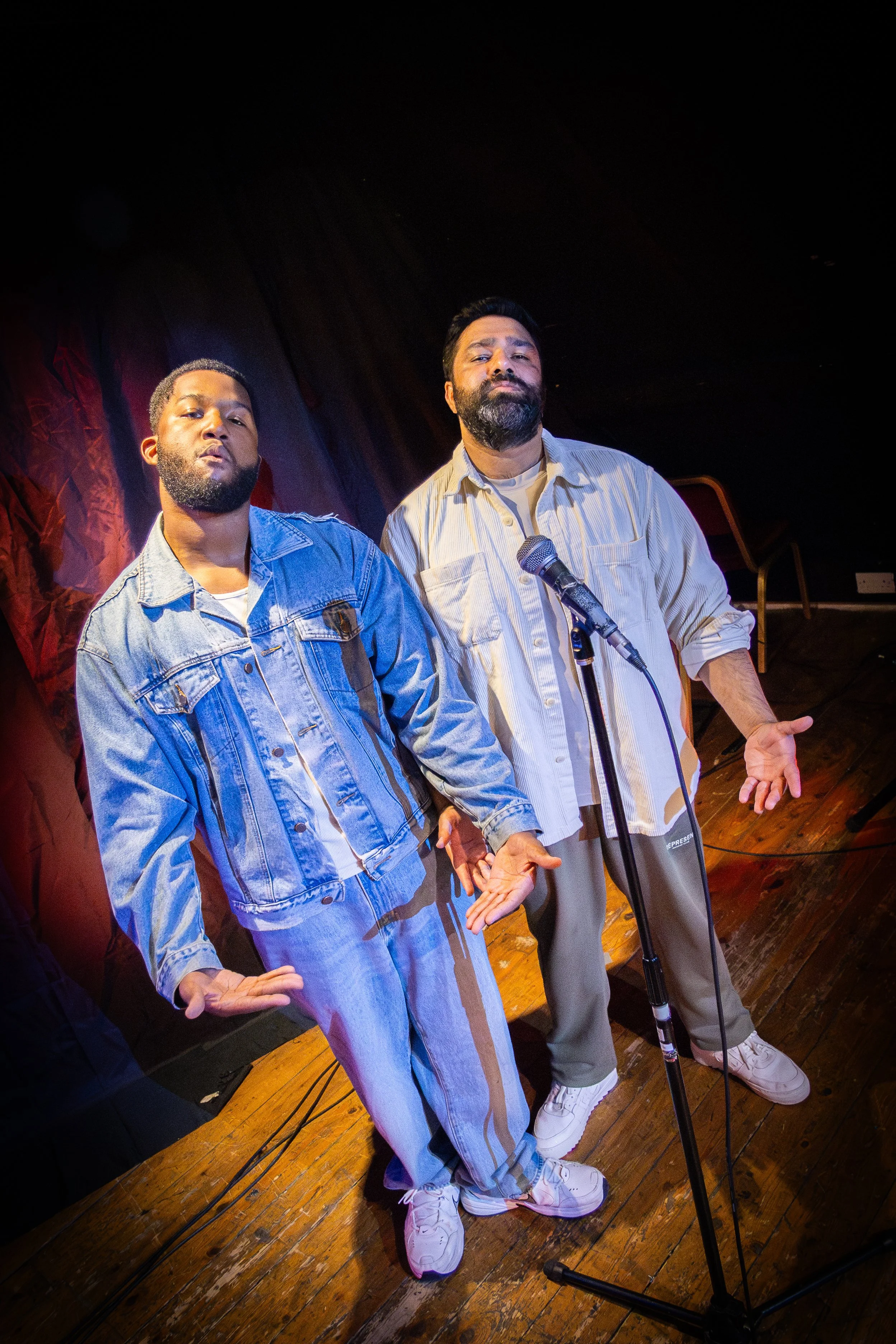 Two men standing together on a stage holding hands, with a microphone in front of them, in front of a black curtain and wooden floor.